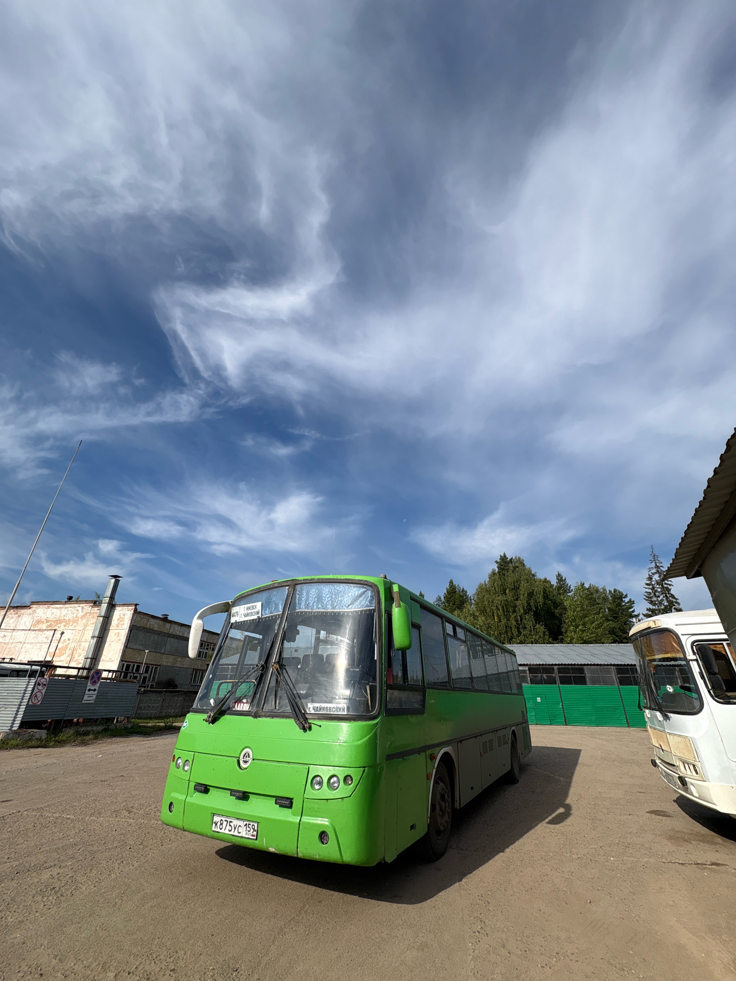 A bright green bus is parked on a dirt lot in front of an industrial building under a cloudy blue sky. The bus, which appears to be in good condition, has its headlights on and the windshield is clean. A white bus is parked to the right of the green bus. The building behind the green bus is a light-colored, multi-story structure with large windows. Trees are visible in the background. A sign on the front of the green bus reads "Г. ИЖЕВСК - Г. ЧАЙКОВСКИЙ". The license plate on the green bus reads "К875УС 159". The location is Noviy, Russia.