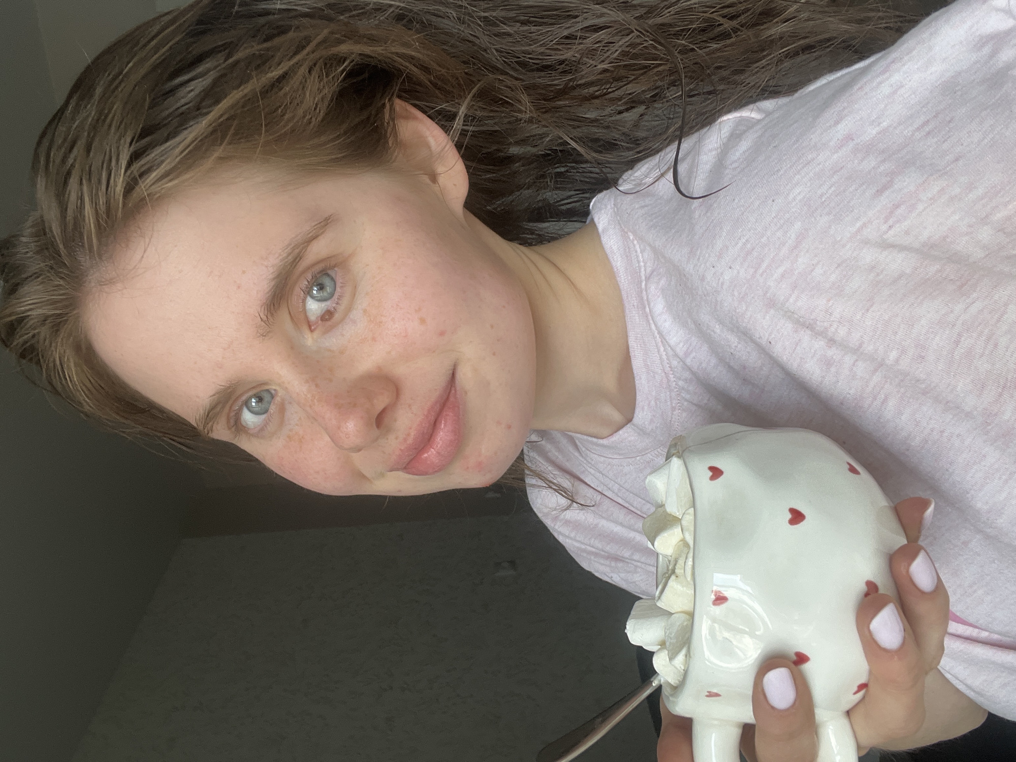 In Chaikovsky, Russia, a young woman with fair skin, freckles, and light blue eyes is holding a white mug filled with marshmallows. Her hair is long and brown, and she is wearing a light pink t-shirt. The mug is decorated with small red hearts. The background is out of focus, showing a dark wall. The lighting appears to be natural, coming from the left.