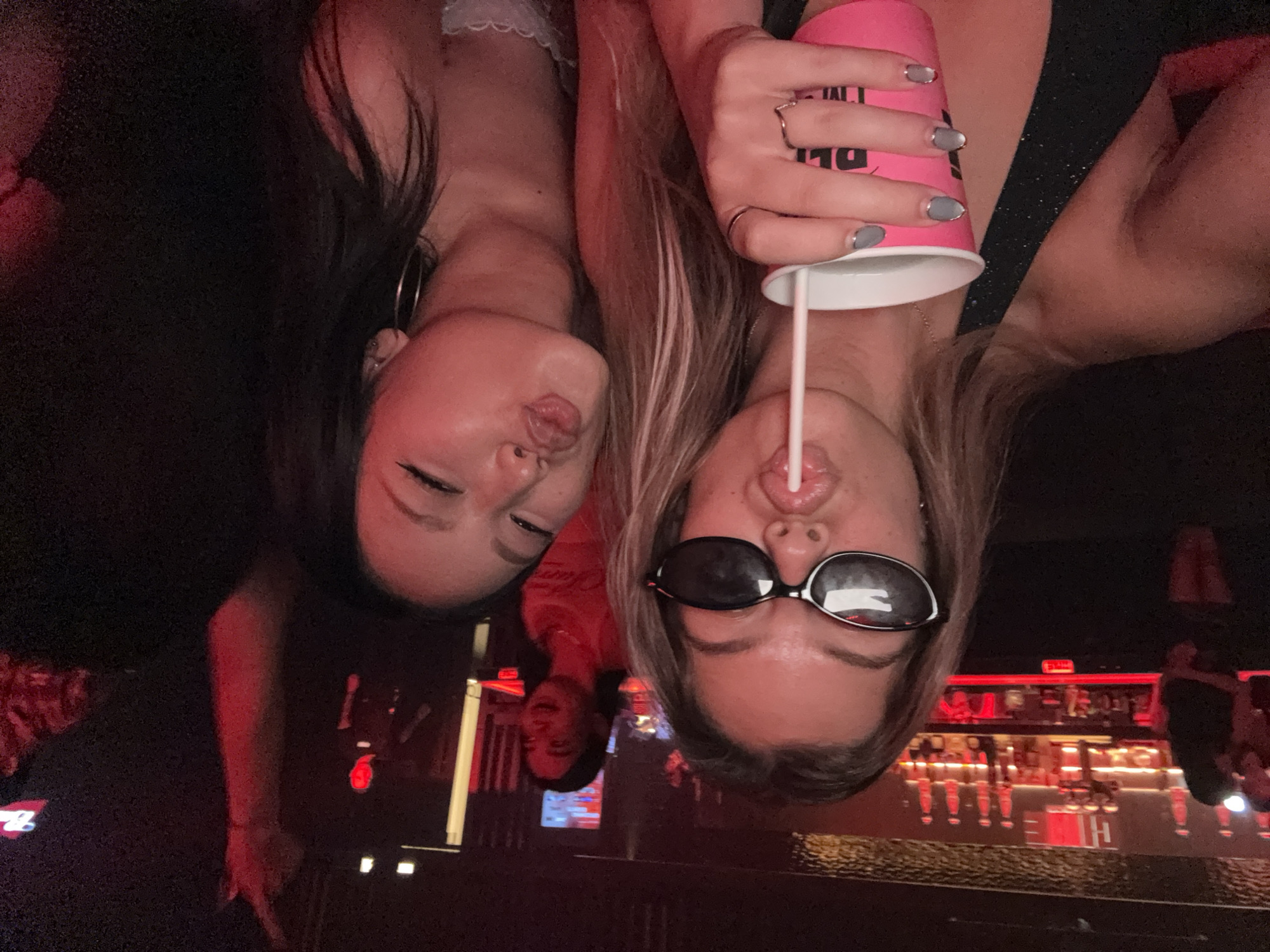 Two women are posing for a selfie in front of a bar. The woman on the left has long dark hair and is wearing a hoop earring. The woman on the right is wearing sunglasses and has a pink cup with a straw in her mouth. Behind them is a bar with alcohol bottles and beer taps, illuminated with red light. The location is in the city of Thủ Đức, Vietnam. A person is visible behind the women, standing at the bar.