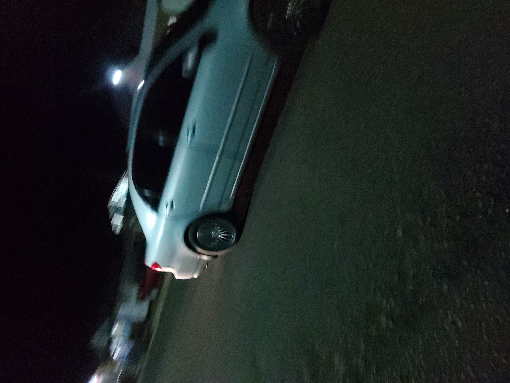 A light-colored sedan is captured in motion at night on a dark asphalt road. The car's rear taillight is visible and illuminated, casting a red glow. The image appears to be taken from a moving vehicle, resulting in motion blur throughout the scene. In the background, out-of-focus shapes suggest buildings or structures, and a distant streetlight emits a bright white light. The overall environment is dark, indicating nighttime. The scene is consistent with a street in Jalingo, Nigeria, where such lighting and road conditions are common at night.