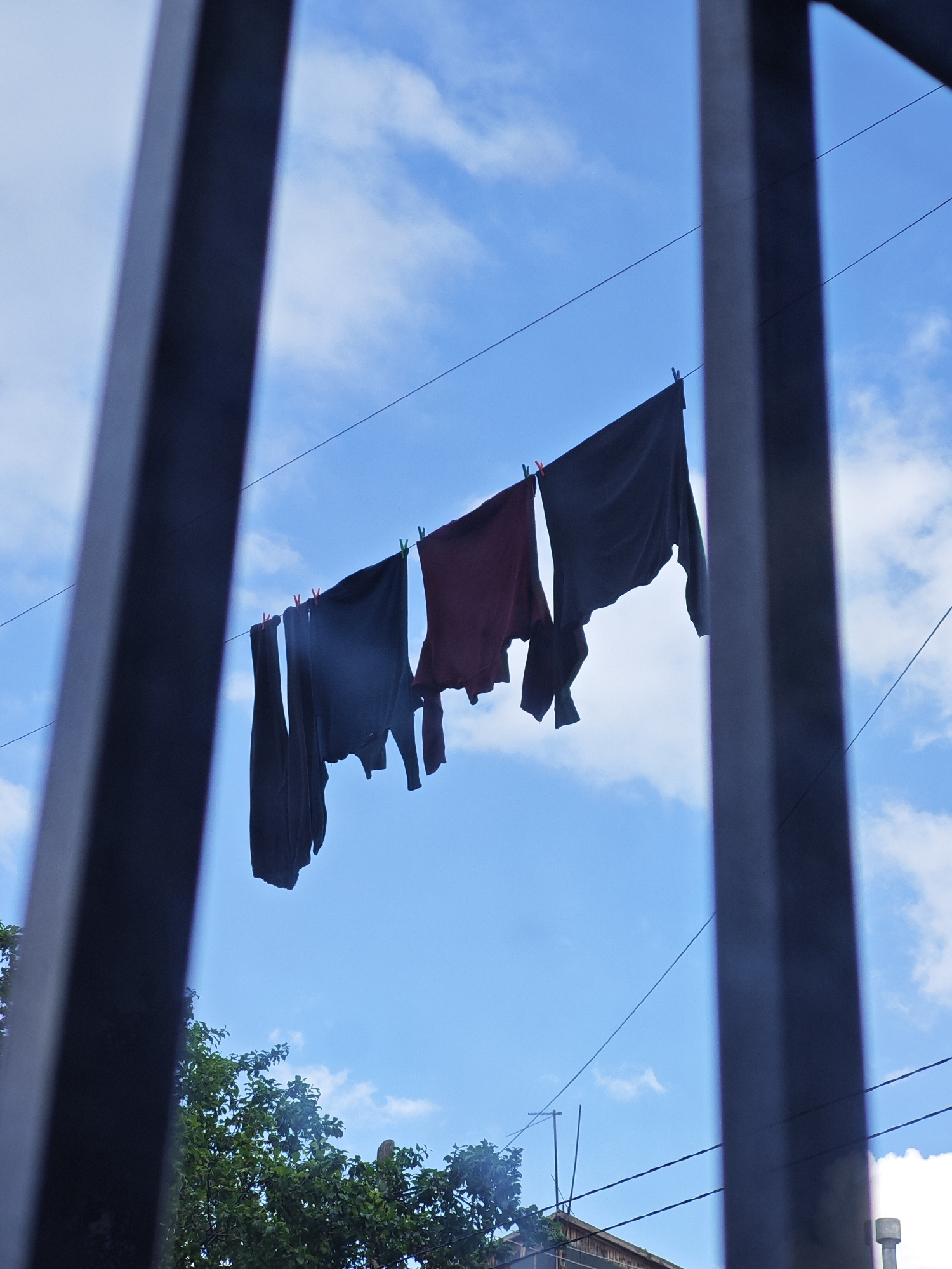 The image, taken in Vanadzor, Armenia, depicts clothing hanging on a clothesline against a blue sky with scattered white clouds. The clothesline stretches across the frame, supported by unseen points. Several garments, including dark-colored shirts and a maroon shirt, are secured to the line with clothespins. The foreground is partially obscured by dark, vertical bars, suggesting the photo was taken through a fence or window. In the lower portion of the image, the tops of green trees and the roofline of a building are visible, indicating an outdoor urban or semi-urban setting. The lighting suggests it is daytime.