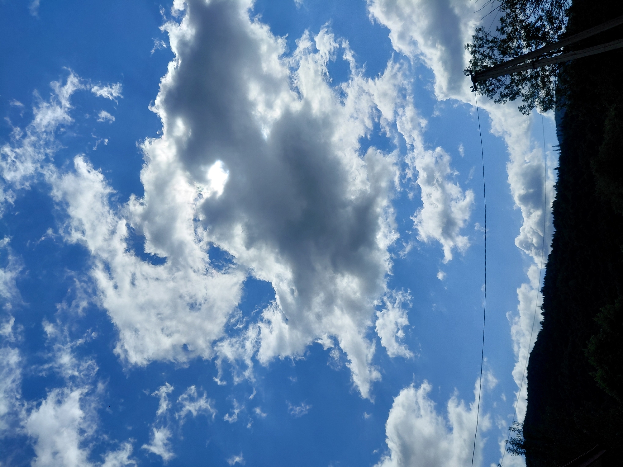 A bright blue sky filled with scattered white and grey clouds is visible above a dark, tree-covered hillside. A telephone wire stretches horizontally across the lower portion of the sky. In the lower right corner, a silhouette of a tree and part of a utility pole are visible against the bright sky. The image appears to be taken in Chernyk, Ukraine, based on the context provided.