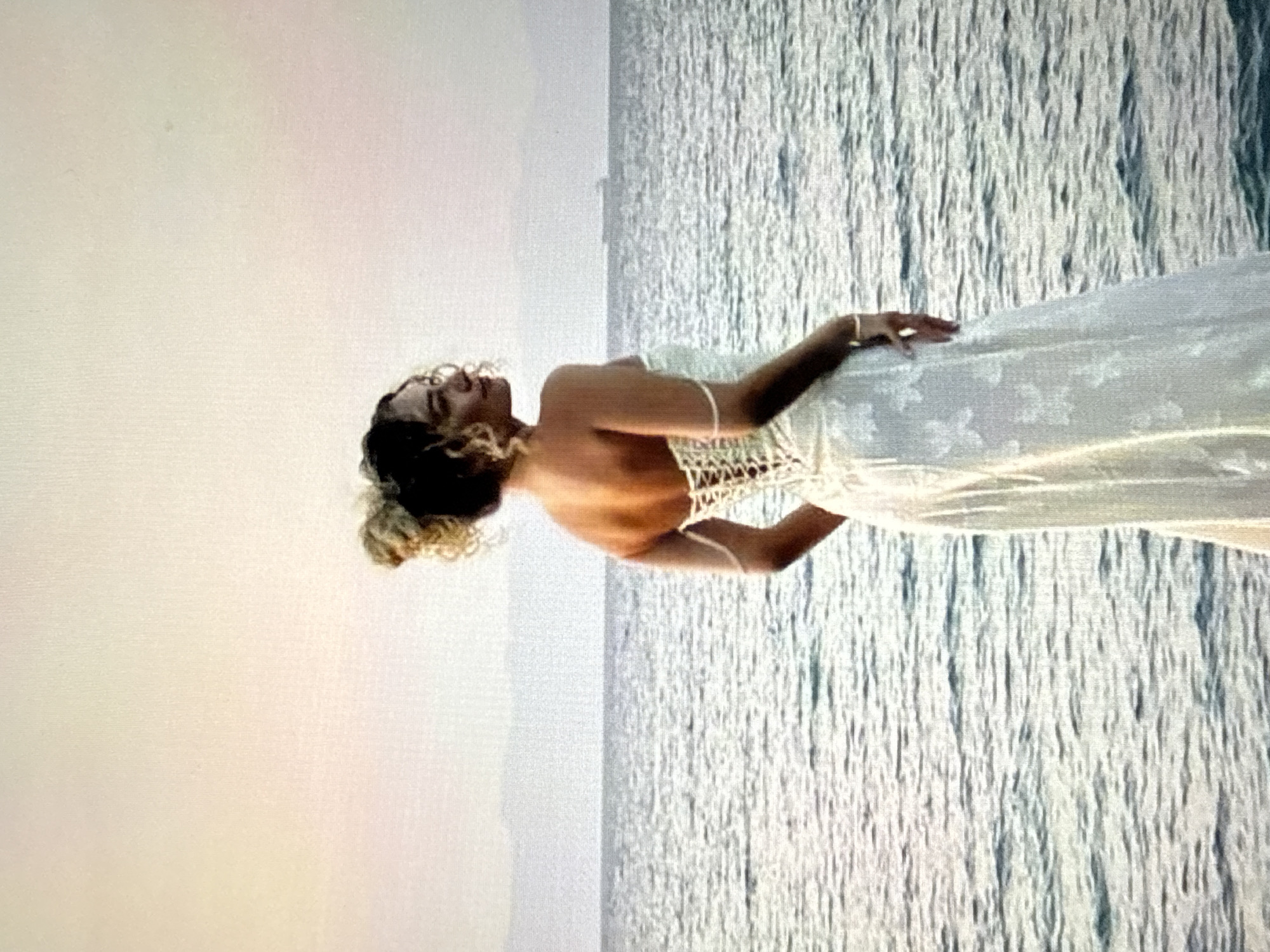 A young woman with curly blonde and brown hair, styled in a bun, stands facing away from the camera, looking to her right. She is wearing a white corset-style dress with delicate white floral embroidery on the skirt. Her back is visible, with the lace-up detailing of the corset prominently displayed. She is positioned on what appears to be a rocky shoreline, with the sea stretching out before her. The water is rippling, and a distant ship or large boat is visible on the horizon. The sky is a soft blend of pale orange and pink, suggesting either sunrise or sunset. The overall scene is serene and picturesque, set against a natural backdrop. The location is not in the city of Găvănoasa, Moldova.