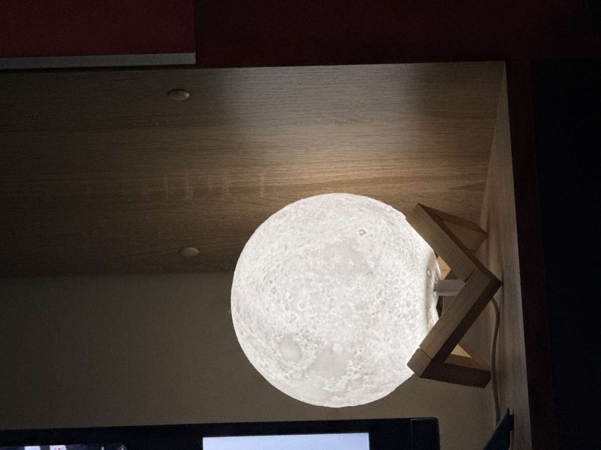 The image shows a dimly lit interior scene. A round, textured lamp resembling the moon is prominently displayed, supported by a wooden stand consisting of interlocking angular pieces. The lamp emits a bright, soft glow, illuminating the surrounding area.
Above the lamp, there's a wooden shelf or cabinet surface with a smooth, light-colored finish. A small, circular recessed light fixture is visible on the underside of the shelf. To the right of the lamp, a dark, solid-colored wall or panel creates a strong contrast.
In the lower part of the image, the top edge of a television screen or monitor is visible, suggesting the presence of electronic equipment in the room. The overall environment appears to be a modern or contemporary interior, likely a bedroom or living space. The scene's location is unknown.