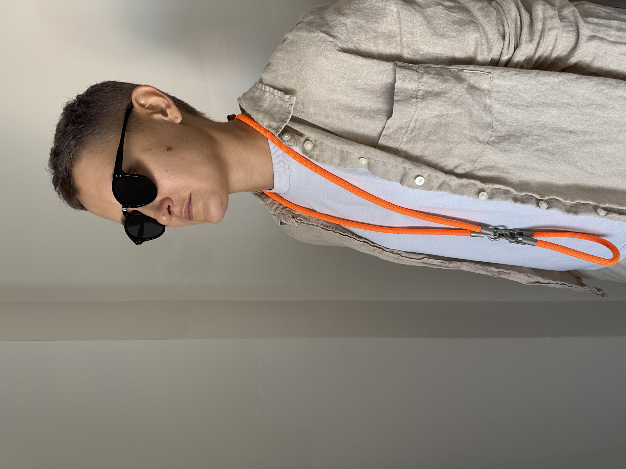 A person with short hair and sunglasses is standing in front of a wall. They are wearing a white t-shirt and a light-colored button-up shirt. An orange cord necklace is visible around their neck. The background is a plain wall, and there is a faint vertical line suggesting a corner or a seam. There are no identifiable landmarks or clues to suggest a specific location in Nha Trang Ward, Vietnam.
