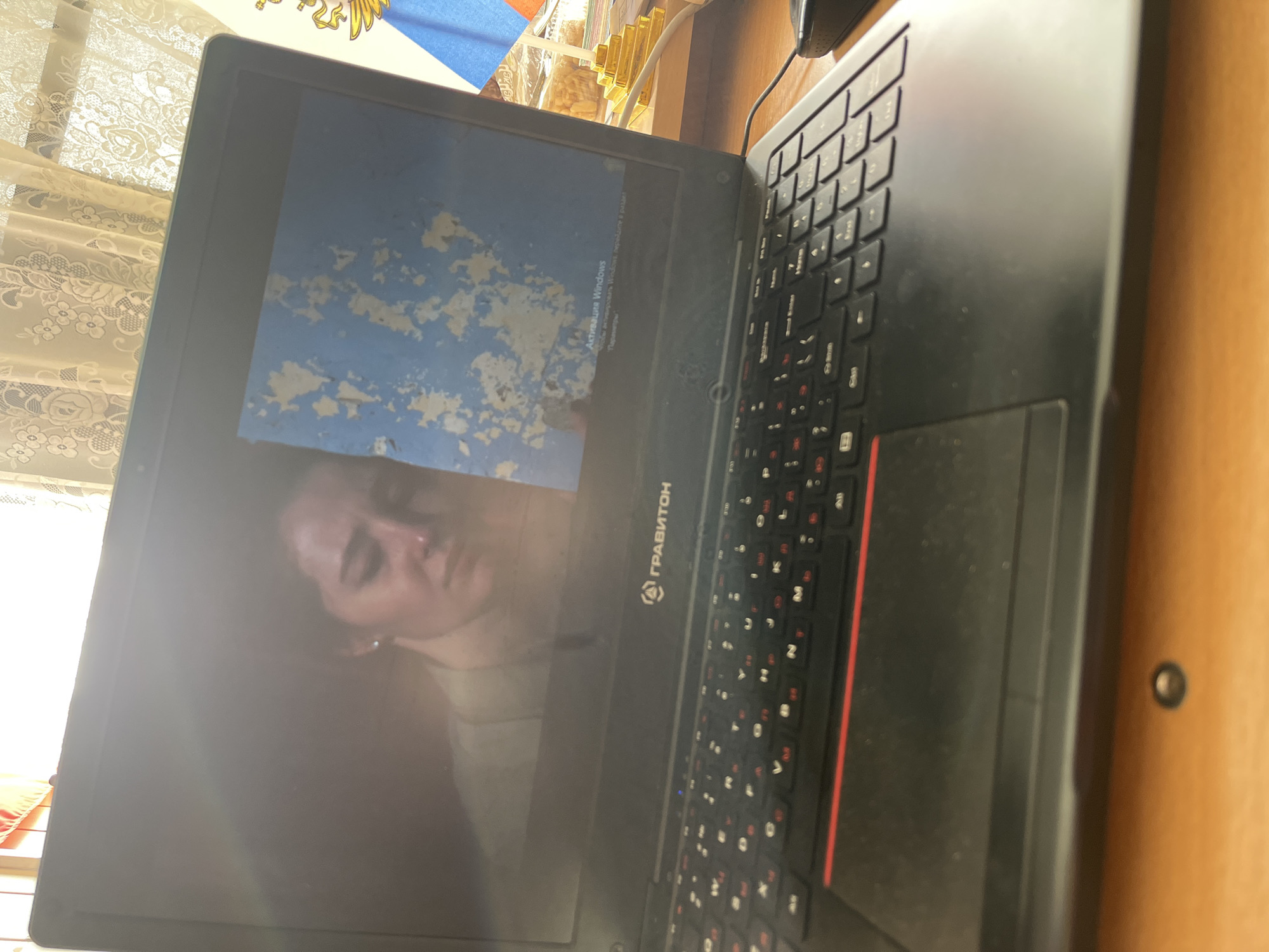On a wooden surface, a partially open laptop displays a reflection of a person. The laptop keyboard is visible, with red accents. A Russian flag can be seen in the background. The laptop brand name "ГРАВИТОН" is visible on the device. The screen shows a message about Windows activation. The location appears to be indoors, possibly a home. It is not possible to establish the specific city as Chaikovsky, Russia, from this image alone.