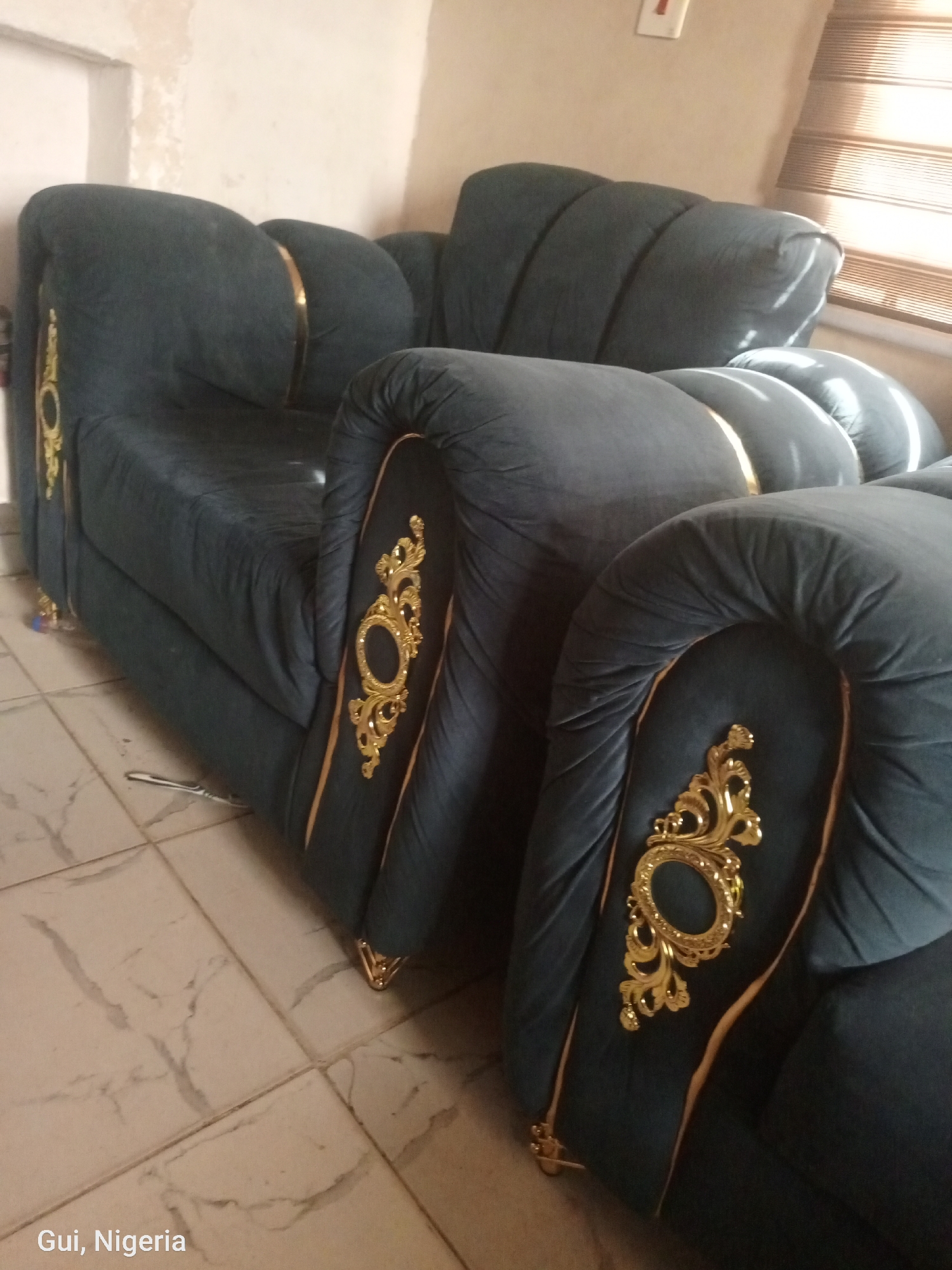 The image shows a dark teal sofa with gold trim and ornate gold detailing on the arms. The sofa is made of a plush fabric and appears to be in good condition. The sofa is situated on a tiled floor. In the background, there is a wall and a window with blinds. The scene is indoors, likely in a residential setting. The text "Gui, Nigeria" is visible in the bottom left corner, suggesting the location.