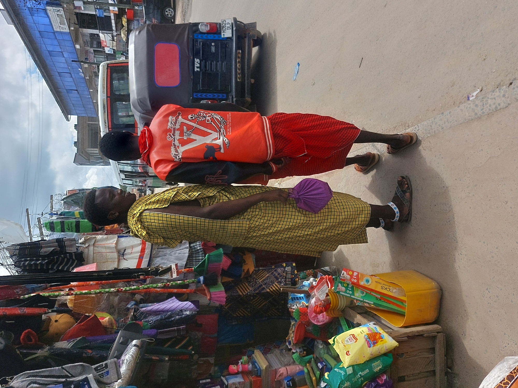The image shows a street scene in Dar es-Salaam, Tanzania. Two people are standing next to a roadside vendor. One person is wearing an orange shirt and red shorts, and the other person is wearing a yellow dress and carrying a purple fan. The vendor stall is filled with various items, including toiletries, small toys, and household goods. Behind them is an auto rickshaw, also known as a "tuk-tuk" or "bajaji", with license plate number MC825 DLP. The area has buildings on the left side.
