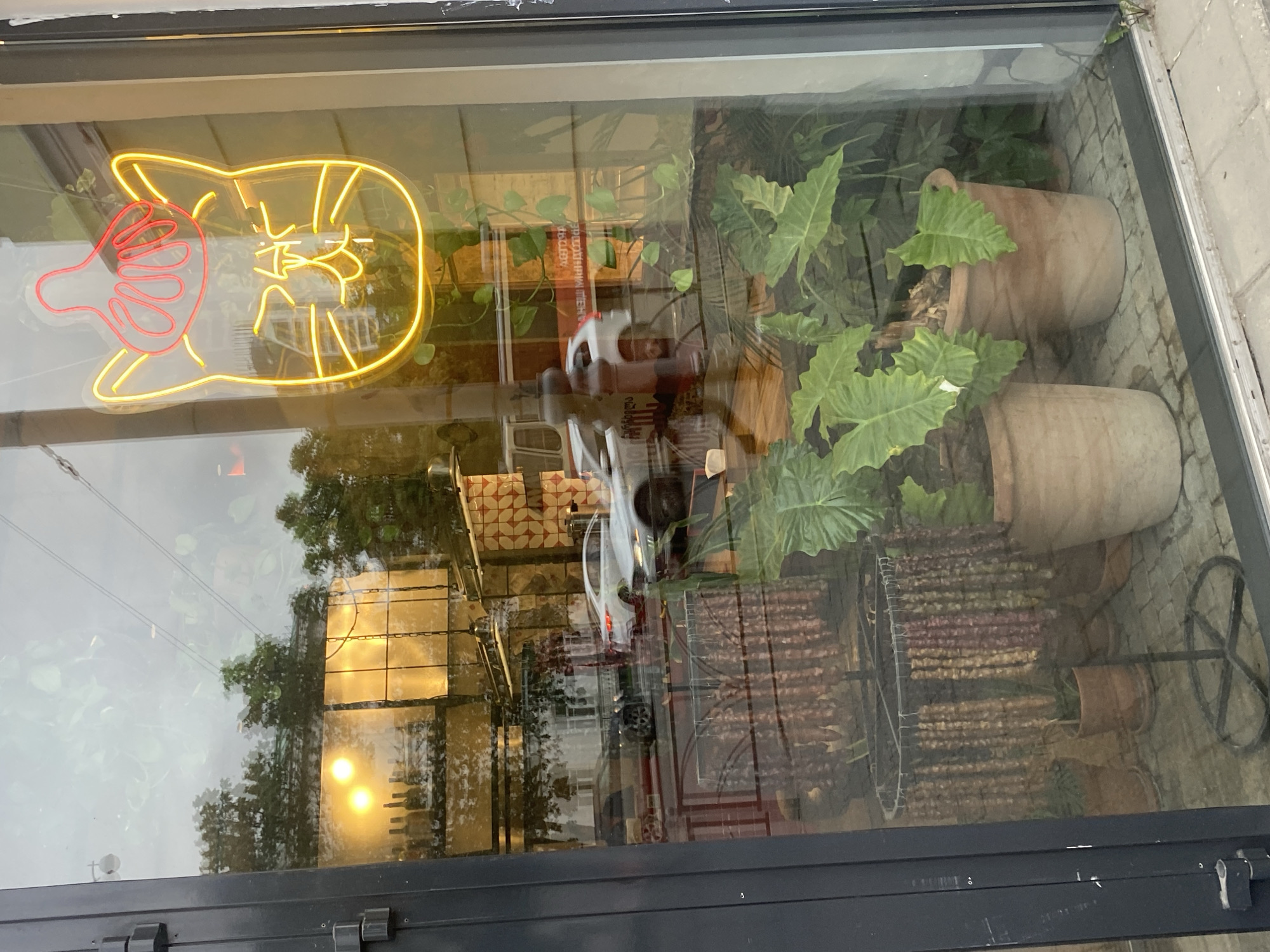 The image is a reflection in a window, likely of a restaurant or shop, in Yekaterinburg, Russia. The window displays a neon sign with a pig, and vertical skewers on metal racks, suggesting a grilling or barbecue theme. Two large terracotta pots containing plants are also visible. The reflection in the glass shows a street scene with buildings, including one with a grid pattern. A white vehicle is reflected in the glass, along with pedestrians, adding to the urban setting. The door frame is dark colored.