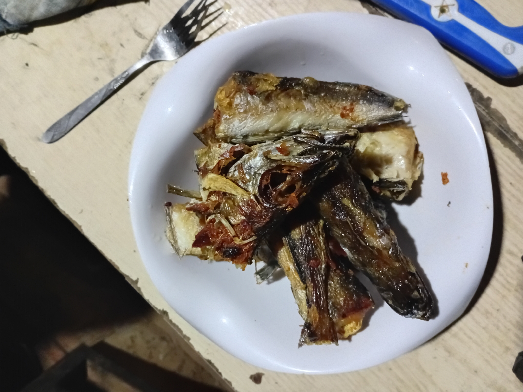 A white plate of fried fish sits on a worn, light-colored wooden surface. A metal fork lies to the left of the plate, and a blue and white object is visible on the right. The scene appears to be indoors. There is no specific evidence indicating that this image was taken in the city of Ikorodu, Nigeria.