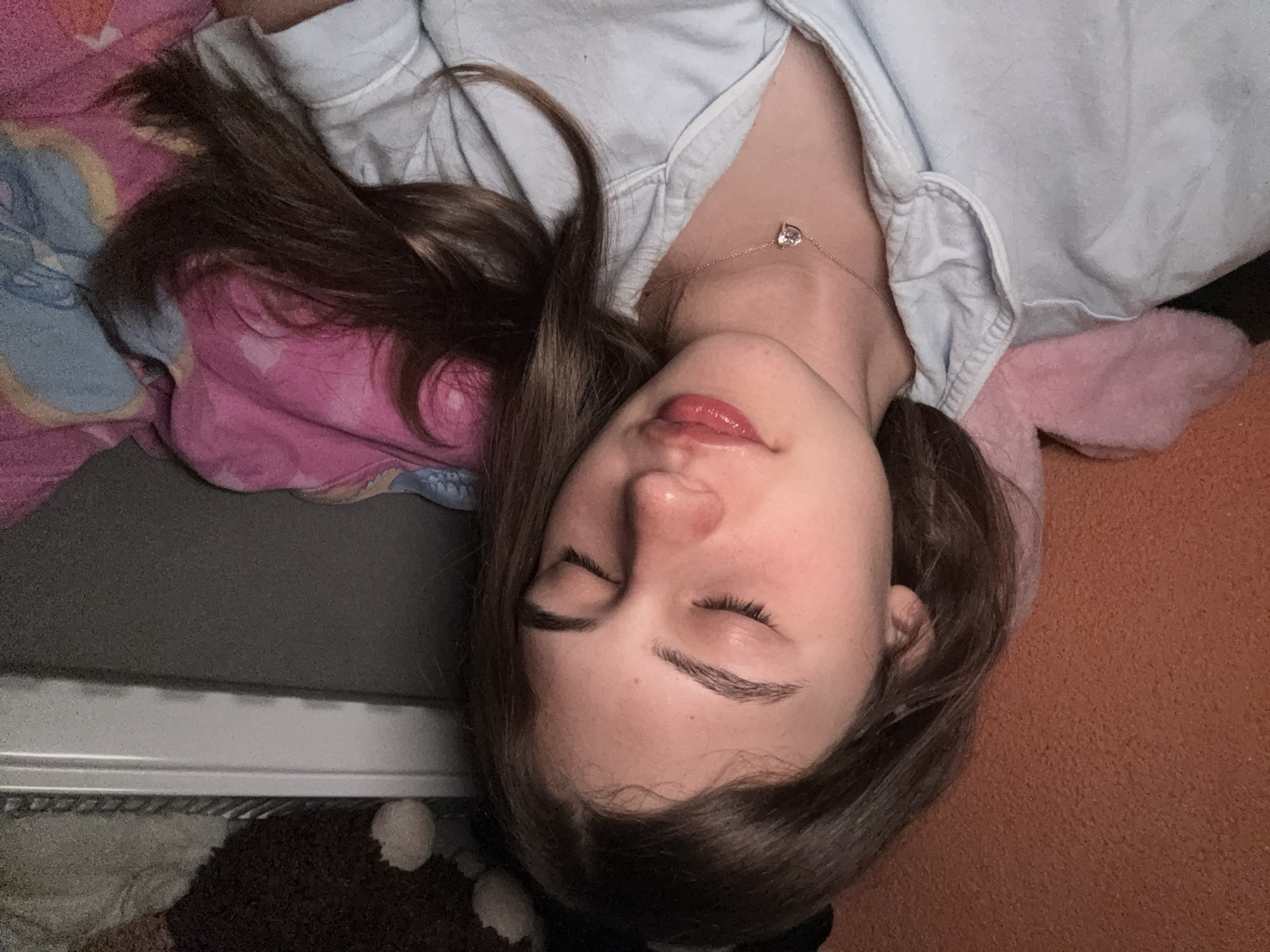 A young woman lies on her back, seemingly asleep. She has medium-length brown hair and closed eyes. She wears a light blue shirt and a silver necklace with a heart pendant. She is on a bed with pink and blue patterned bedding and a pink pillow. The background includes a gray surface and an orange surface. The location is not possible to determine from the image.