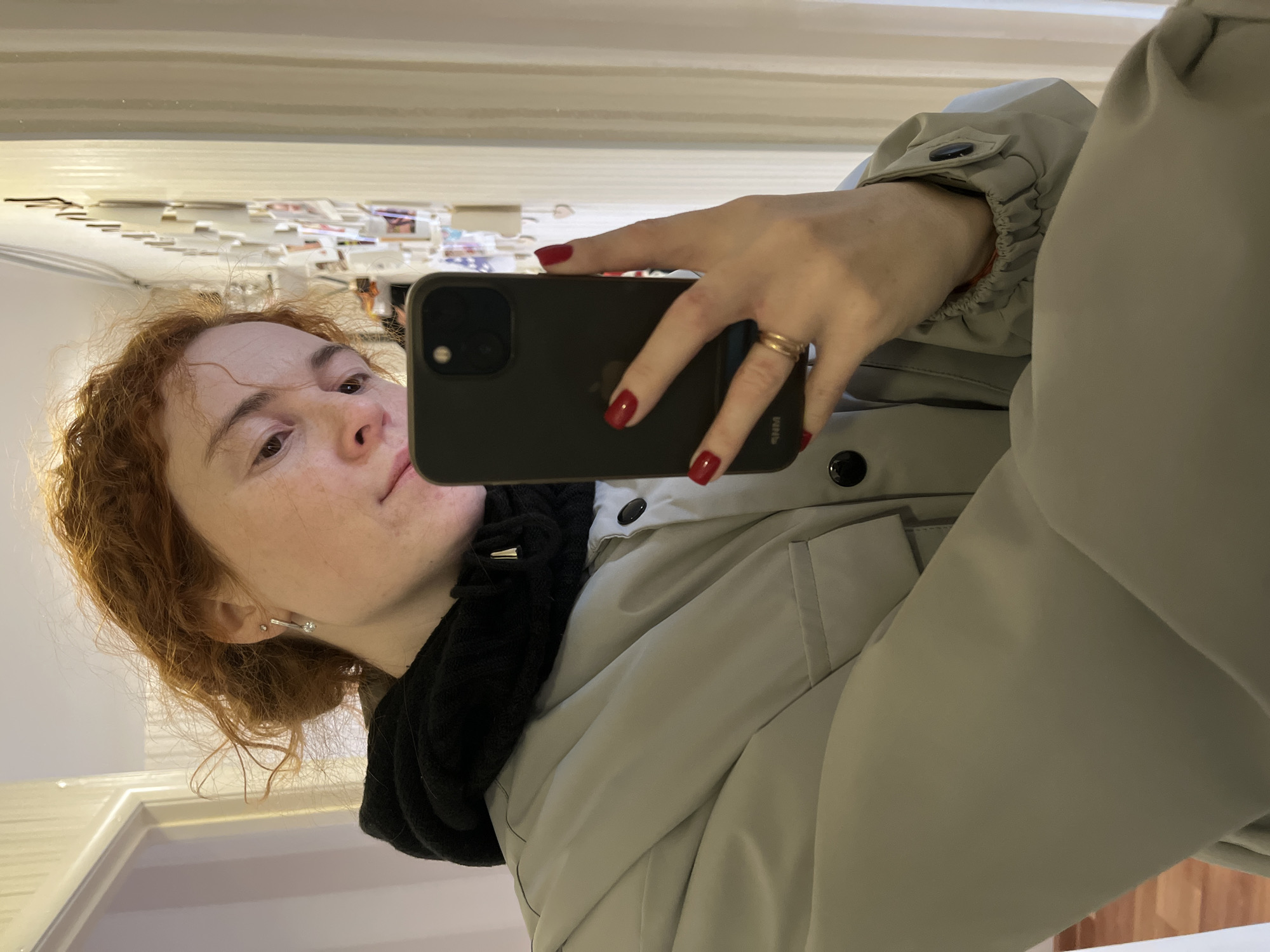 A red-haired woman is taking a selfie in front of a mirror. She is holding a black phone with red nail polish on her nails. She is wearing a light-colored jacket and a black scarf. The reflection in the mirror shows a white wall with various items attached. The image was taken in the city of Chaikovsky, Russia.