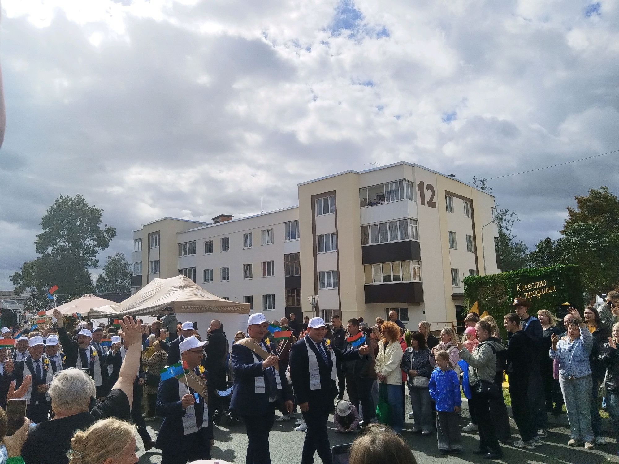 The image shows a street scene with a group of people, many wearing white baseball caps and some holding flags. A building with the number '12' visible on its facade stands in the background. The building appears to be a multi-story residential or office building. There are tents set up on the street, suggesting an event or market. In the background, there is a green wall with text in Russian that translates to "Quality and Traditions." This text, along with the architectural style of the building and the overall atmosphere, suggests the location is likely a public space in Brest, Belarus, possibly during a festival or public gathering. The people are diverse in age and gender, and many are looking towards the center of the frame, with some waving or holding up phones, indicating they are observing or participating in an event. The sky is overcast with some patches of blue visible.