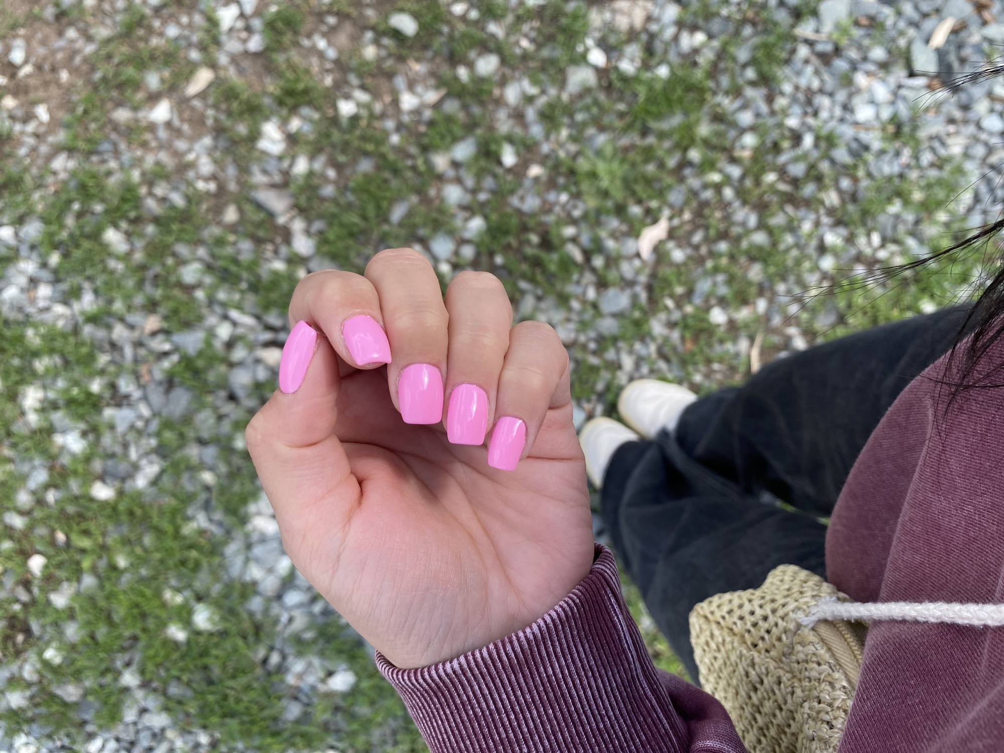 The image displays a close-up, high-angle view of a person's hand and lower body set against an outdoor ground, located in Chaikovsky, Russia. The primary focus is a hand, likely belonging to a female, showcasing short, neatly manicured nails painted in a vibrant, glossy bubblegum pink color. The fingers are slightly curved, and the palm is partially visible. The person is wearing a maroon or burgundy long-sleeved top or sweatshirt, with the ribbed cuff visible around the wrist.
In the background, below the hand and arm, are the person's legs, dressed in loose-fitting dark (possibly black) pants and white shoes. A light brown or beige woven bag, resembling straw or crochet material, is visible near the person's hip. The ground beneath consists of a scattered mix of small, light-colored gravel stones and patches of green grass, suggesting an unpaved path or natural area. The lighting indicates it is daytime, with soft light and no prominent shadows. There is no visible text. The scene conveys a casual moment, possibly taken while the person is outdoors, highlighting their new manicure.