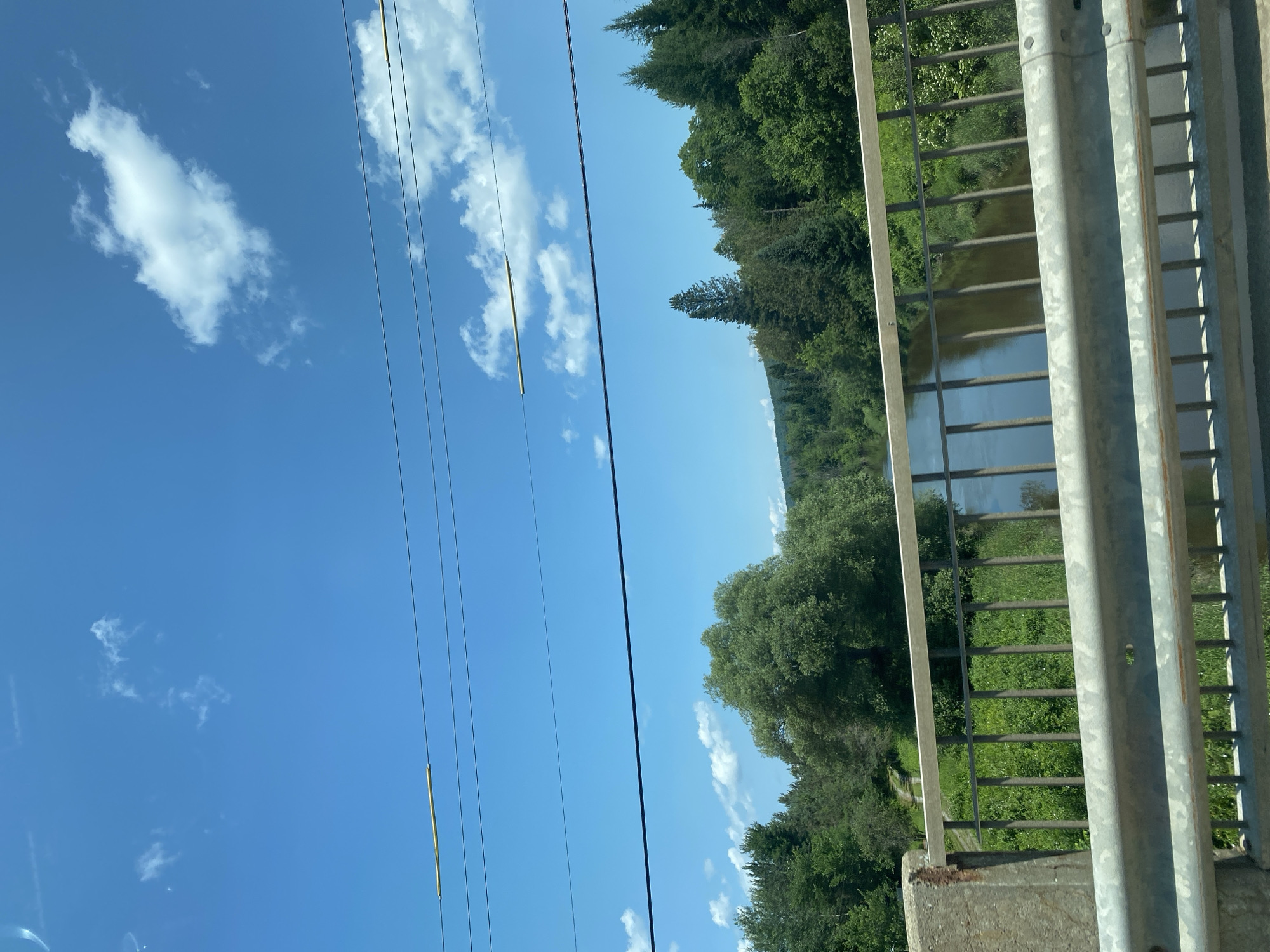 The image shows a view from a bridge over a river, with lush green trees and vegetation lining the banks. The sky is a bright blue with scattered white clouds, and several power lines run horizontally across the top of the frame. The bridge itself has a concrete support on the left and a metal railing. The river appears calm, reflecting the surrounding greenery and sky. The scene suggests a natural, possibly rural or park-like setting within Saint-André-Avellin, Canada.