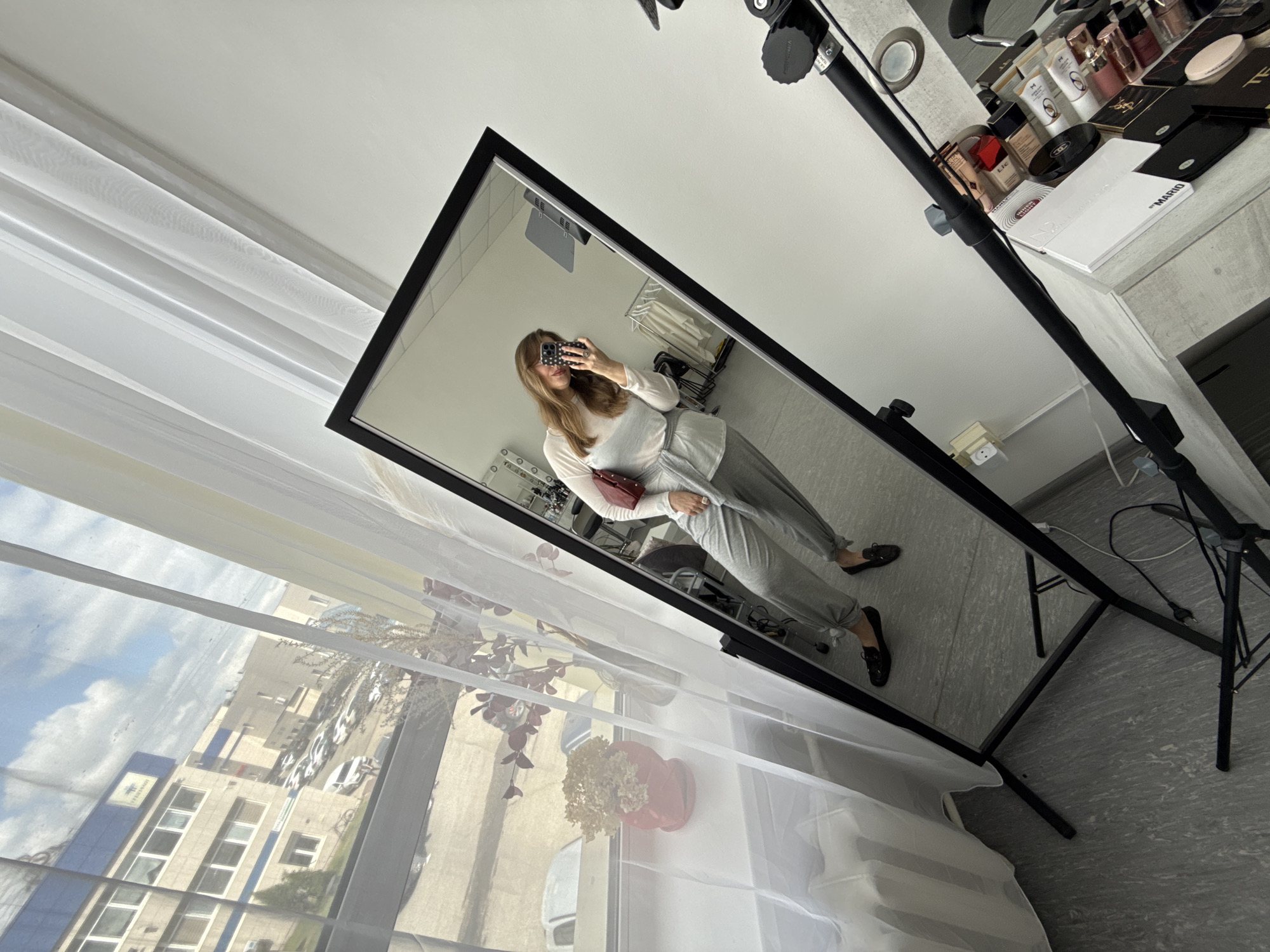 An indoor mirror selfie captures a woman taking a picture of her reflection, with a window and outdoor scene visible to the left. The entire image is significantly tilted clockwise.
In the foreground, translucent white curtains hang over a window. Beyond the curtains, a daytime urban or suburban environment is visible under a sky with scattered clouds. Buildings, including a prominent blue and white structure with a partial logo, along with parked cars and some trees, are seen outside. On the window sill, there's a red pot containing dried flowers.
The central subject is a tall, black-framed full-length mirror reflecting the woman. She has long, light brown hair and is wearing a light-colored, long-sleeved top, grey pants tied at the ankles, and dark flat shoes. She holds a smartphone with a patterned case to take the selfie, and a small red handbag is in her other hand.
To the right of the mirror, a black stand, possibly for lighting or equipment, extends upwards. Behind this stand, on a surface like a dresser or table, an array of makeup and beauty products is neatly arranged. Visible brands include "CHANEL," "YSL," "TOM FORD," and "MARIO." The room features light grey speckled flooring and white walls. In the mirror's reflection, a clothing rack with light garments is visible behind the woman. The setting suggests a personal space, such as a dressing room or studio, located in Izhevsk, Russia.