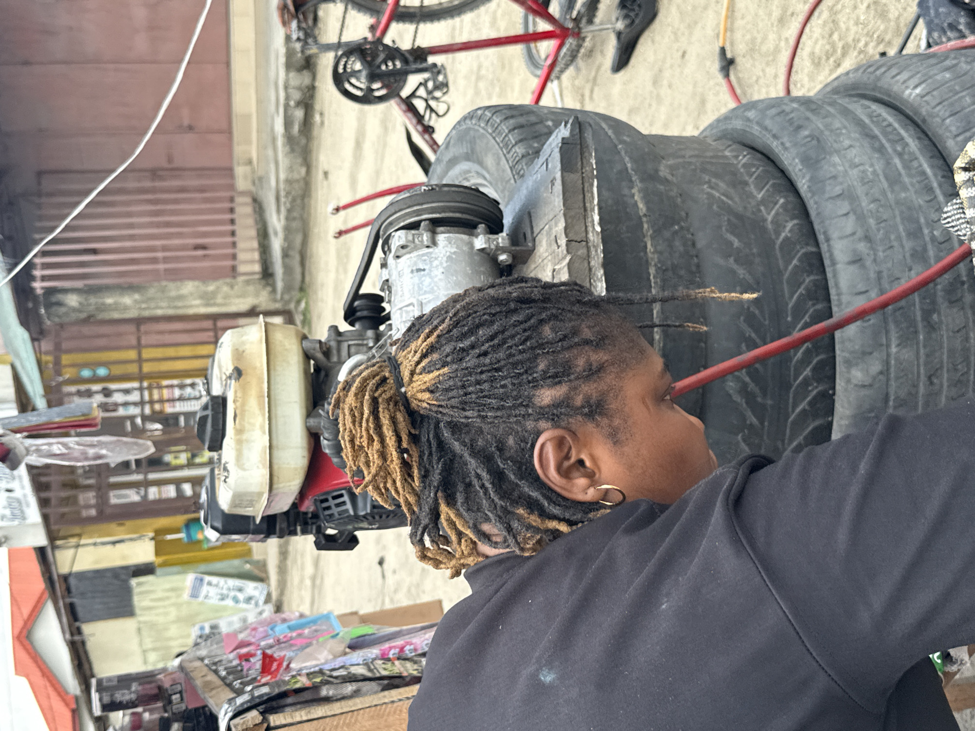 A person with braided hair, wearing a black sweater, is near an engine, possibly doing maintenance. Several stacked car tires are adjacent to the engine. A bicycle, predominantly red, is visible in the background. In the lower portion of the frame, there are various objects, possibly tools or supplies. The scene appears to be outdoors in a semi-urban setting, likely Ado, Nigeria. The background includes walls and possibly commercial or residential structures.