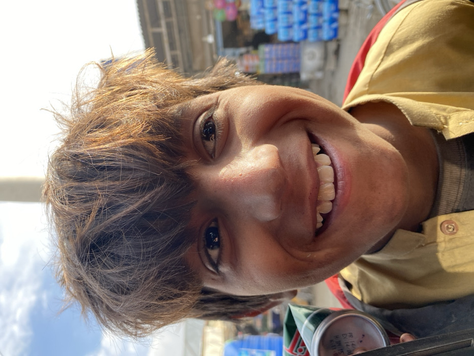 A young male subject, appearing to be a child or adolescent, is centered in a close-up, outdoor portrait taken in Kandahar, Afghanistan. He has a broad smile, revealing white teeth, and looks directly forward. His hair is dark brown with lighter, possibly auburn, tones, styled in a shaggy manner that covers his forehead. The subject's skin tone is olive, with some redness observed on the nose and cheeks. He wears a light-colored, possibly tan or mustard-yellow, collared garment, open at the neck, over a darker, possibly grey, undershirt. A red stripe or part of another garment is visible on his right shoulder. In the lower-left corner, a portion of a metallic green and silver can, bearing white text including "EXP: 14:00," is partially visible, held by the subject.
The background, blurred, depicts an urban or market environment under a clear blue sky with sparse white clouds. To the far left, a light-colored structure, possibly a tent or tarp, is present. Behind the subject's head, a light-colored pole or column extends vertically. To the right, a commercial building with a corrugated metal roof and what appear to be concrete or brick walls is visible. Outside this building, stacks of blue bottled water are prominently displayed, along with colorful balloons (red, pink, blue, green) near an entrance. A bicycle wheel rim is faintly discernible behind the subject's right side. Strong sunlight illuminates the scene, casting highlights on the subject's hair and face.