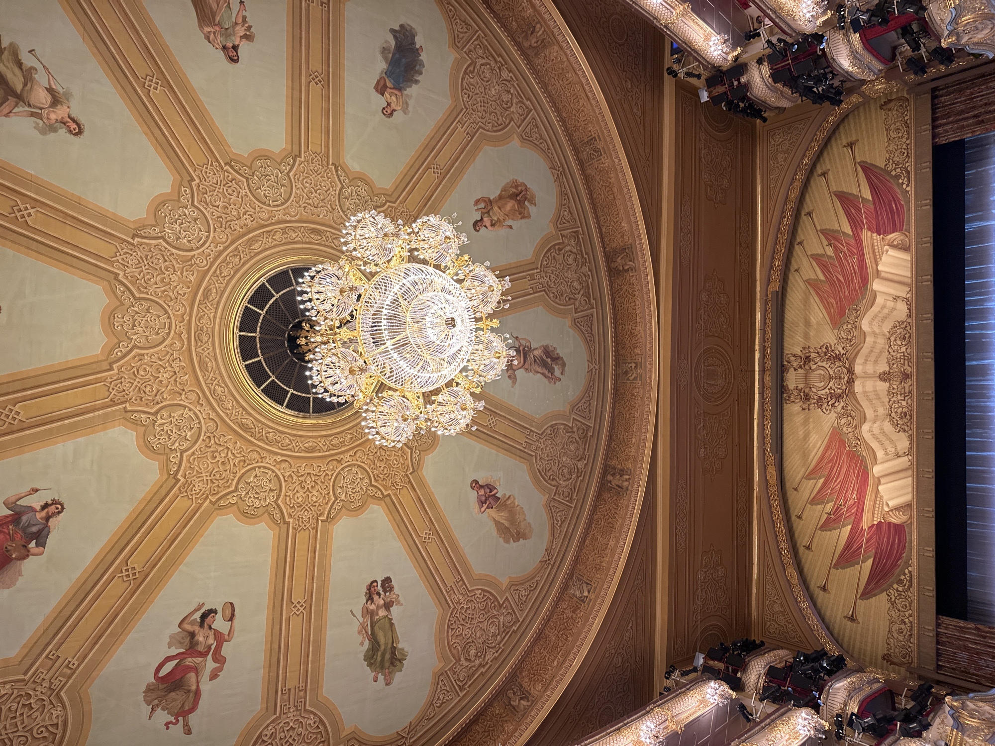 This upward-facing image captures the opulent interior of a grand theatre or opera house, focusing on its magnificent ceiling and a central chandelier. A large, multi-tiered crystal chandelier dominates the center, emitting a brilliant, warm light. It hangs from an elaborate, circular ceiling medallion surrounded by intricate golden scrollwork and radiating decorative beams.
The ceiling sections are adorned with painted murals featuring approximately eight allegorical figures. These figures, predominantly women in classical attire, are depicted holding various artistic or musical instruments and objects, such as a tambourine, a trumpet-like instrument, a painter's palette and brush, or a quill and scroll, suggesting muses of the arts. The ceiling's base color is a muted light green, complemented by the rich gold of the decorative elements and the earthy tones of the figures. A circular, gridded opening is visible directly above the chandelier, possibly a skylight or a ventilation feature.
To the right, a section of a richly ornamented proscenium arch or wall is visible. It features impressive murals with stylized red, feather-like elements, alongside golden trumpets, a lyre, and a scroll displaying musical notation, all contributing to a strong theme of music and performance. Below and along the right side of the image, portions of ornate theatre balconies with golden railings and dark seating are visible, equipped with modern stage lighting fixtures.
The scene conveys a sense of historical grandeur and elegance, typical of a prestigious European performance venue. The artificial lighting suggests an indoor setting, likely in the evening. No discernable text is visible. Given the provided context, this elaborate interior is characteristic of a prominent theatre in Moscow, Russia.