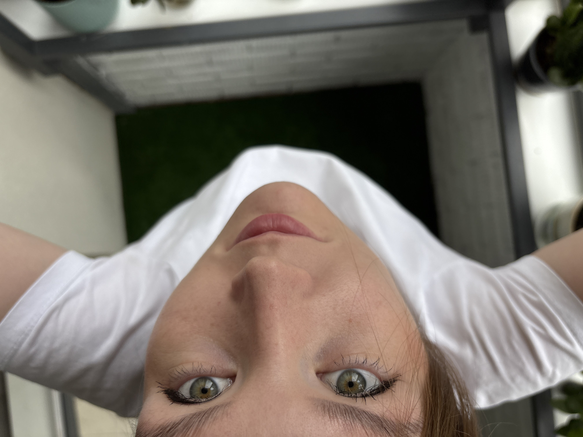 An upward-angle, close-up shot focuses on the face of a young person wearing a white t-shirt. Their head is tilted, and they are looking directly at the camera. The subject has light skin with freckles, prominent eyelashes with mascara, and striking green eyes. Their lips are slightly parted and appear to have a light pink tint. The background is blurred, showing a glimpse of dark green, possibly artificial grass, and a dark gray railing. The overall impression is that the photo was taken outdoors, possibly on a balcony or rooftop. The image does not contain any specific elements that would indicate its location in Izhevsk, Russia.