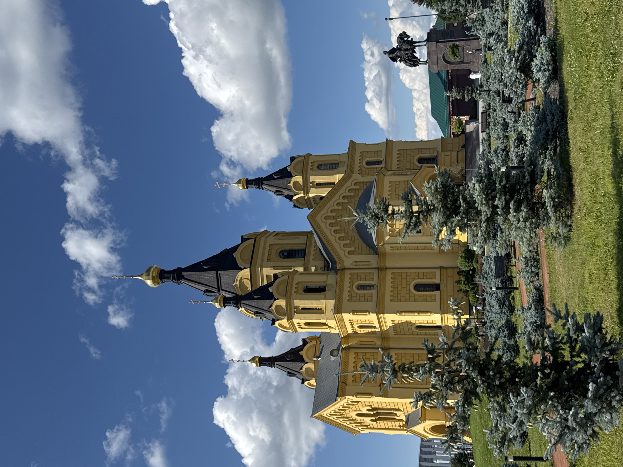 This is a full, eye-level, outdoor, daytime photograph of the Alexander Nevsky Cathedral in Nizhny Novgorod, Russia. The cathedral is a large, bright yellow Orthodox cathedral with black domes and golden crosses. It is situated on a green, grassy hill with several small evergreen trees and bushes in the foreground. In the middle ground, to the right of the cathedral, is a statue of a man on horseback. Behind the statue is a green-roofed building. The sky is bright blue with scattered white clouds. The overall scene is serene and majestic.