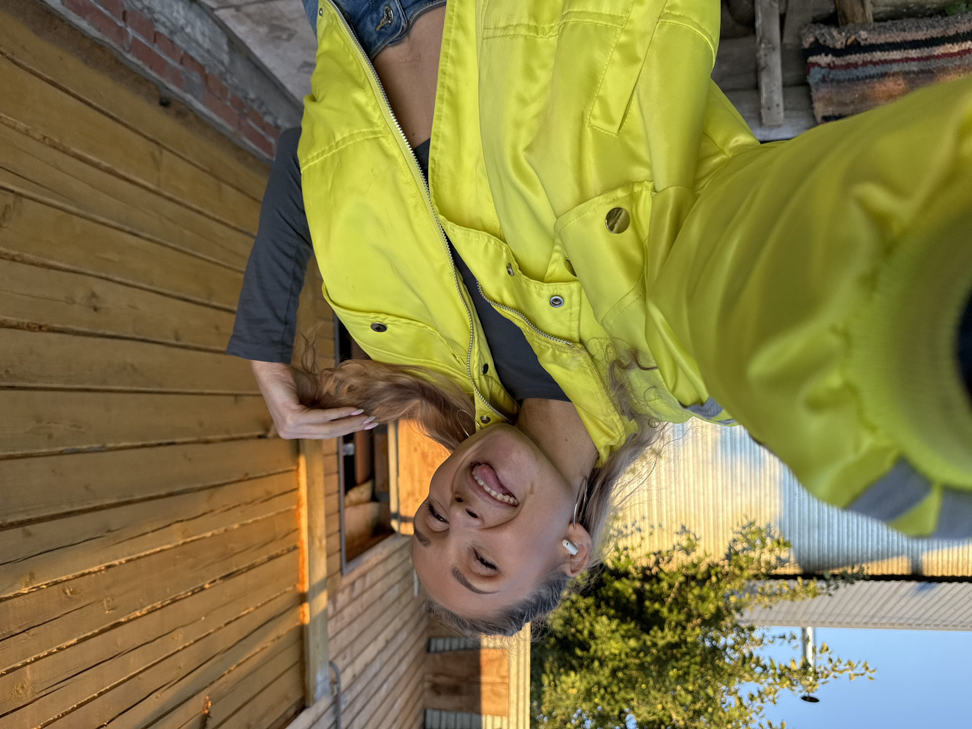 A young woman, likely in her early 20s, with blonde hair is pictured outdoors. She is wearing a bright yellow zip-up jacket over a dark top and denim bottoms. The woman is holding her phone to take a selfie, and her head is tilted back, with her eyes closed and tongue sticking out, suggesting a playful or teasing expression. Large hoop earrings are visible in her ears, and she is wearing white wireless earbuds.
The setting appears to be a backyard or outdoor area, with a wooden fence and a brick wall to her left. Green trees and a bright blue sky are visible in the background. The lighting suggests it is either early morning or late afternoon, with the sun casting a warm glow on the wooden surfaces. The overall mood is casual and lighthearted. There is no visible text in the image.
