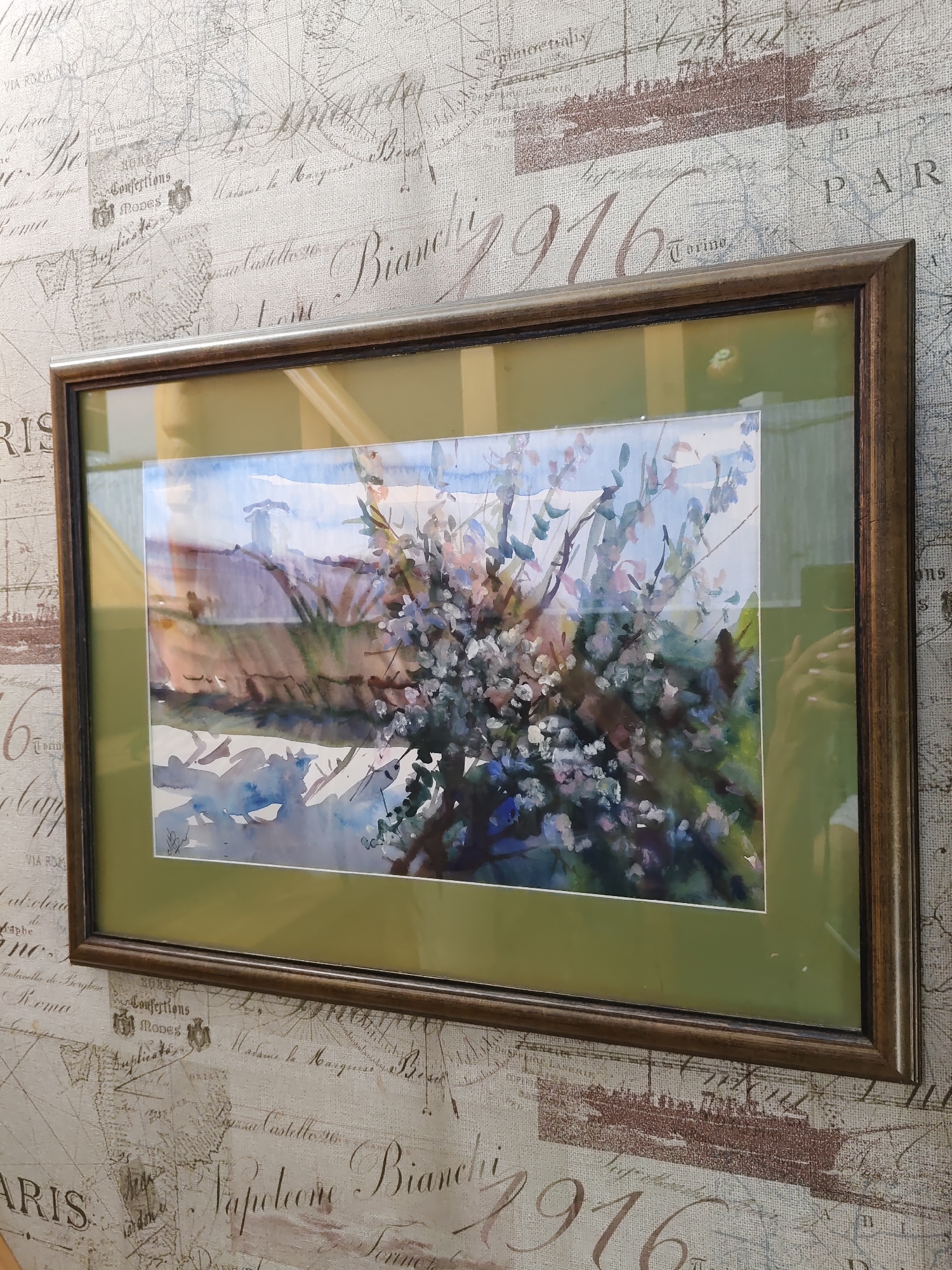 A framed watercolor painting of a flowering tree is displayed against a textured wallpaper featuring a map and cursive text. The painting depicts a tree with delicate pink and white blossoms in the foreground, with a blurred background of green foliage and a pale blue sky. The frame is wooden with a metallic sheen. The wallpaper has a vintage feel with faint map lines and script. The image does not contain any recognizable landmarks or elements that would indicate its location in Rostov-on-Don, Russia.