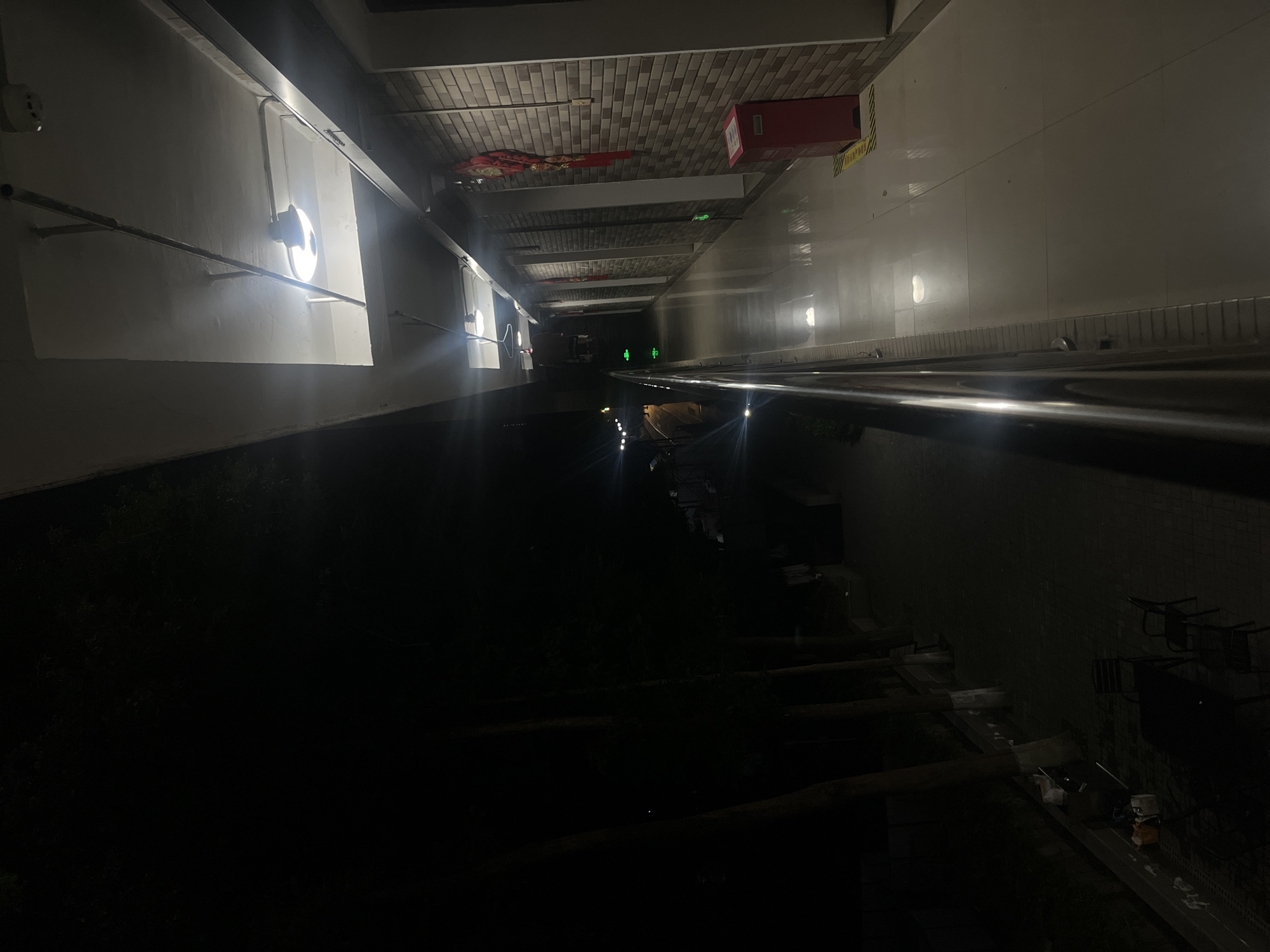 The image captures an elevated open-air corridor at night within Yuzhong District, China. The corridor, situated on the right side of the frame, features a reflective, light-toned tiled floor and a metallic railing to the left. Overhead, bright white circular lights illuminate the space, revealing exposed conduit work on the ceiling. The right wall is faced with textured light-brown tiles resembling brickwork, punctuated by several dark recessed panels and adorned with red traditional Chinese decorations. A red fire extinguisher box rests on a yellow-and-black striped safety mat near the foreground, adjacent to an illuminated green exit sign.
To the left and below the corridor, a dark outdoor environment is visible. This area includes a paved path bordered by multiple dark tree trunks. Faint, distant light sources are discernible further down this lower level. In the bottom-left corner, an outdoor seating arrangement with a dark table and chairs is present, along with miscellaneous items near a low wall. No persons are depicted in the scene.
