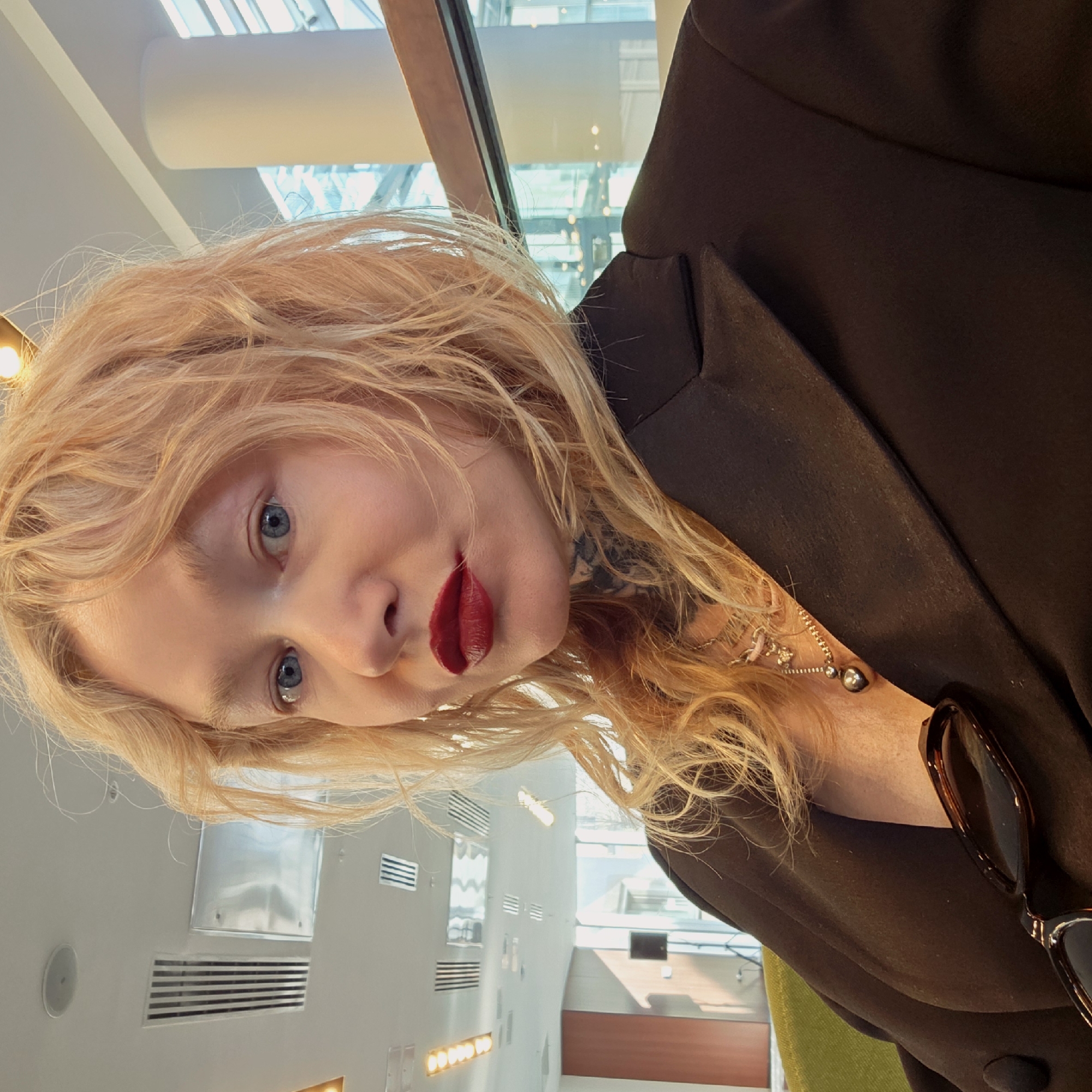 A woman with long, wavy blonde hair, striking blue eyes, and dark red lipstick is depicted in a close-up, slightly upward-angled selfie or portrait. She is wearing a dark, possibly black or very dark brown, blazer or jacket, with what appears to be a dark-colored tattoo visible on her neck/shoulder area beneath her hair. A delicate gold-toned necklace with a small pendant, another necklace with a dark spherical charm, and a small silver-toned earring are visible. A pair of brown, tortoise-shell sunglasses is casually tucked into the lapel of her blazer. Her expression is direct, looking at the camera, with a slight pout.
The setting is indoors, featuring modern architecture with high ceilings and large windows or skylights that let in bright natural light, suggesting it is daytime. The background shows light-colored walls with horizontal ventilation grilles and various rectangular and circular light fixtures embedded in the ceiling and walls. Glimpses of a bright green chair and other structural elements of a contemporary building, possibly an office, airport lounge, or a public space, are visible. The overall impression is bright and spacious. There is no visible text in the image. The location context for this scene is Saint Petersburg, Russia.