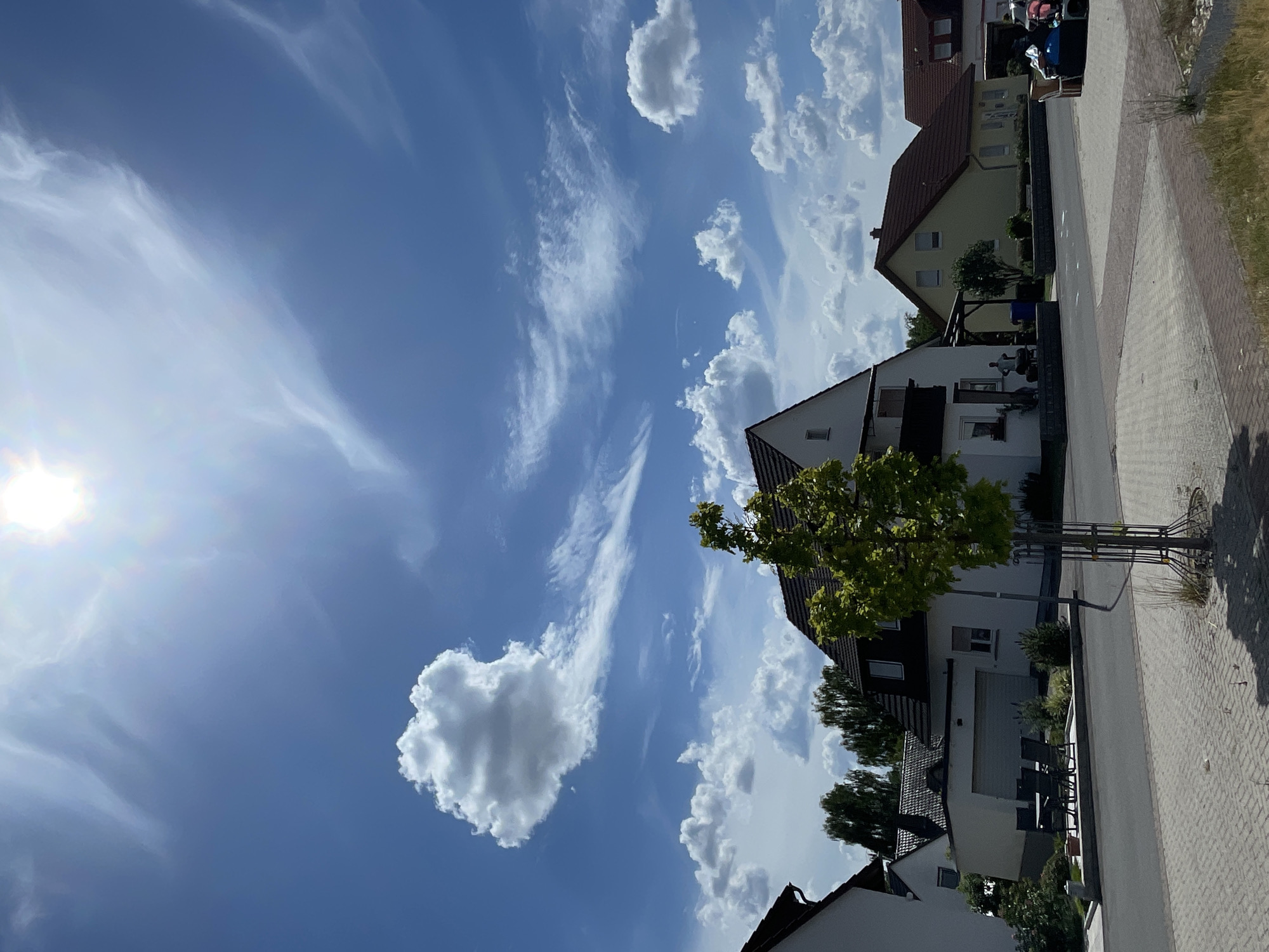 The image depicts a residential street in Röllshausen, Germany, under a bright, sunny sky with scattered clouds. In the foreground, a paved street runs horizontally across the frame, with a brick-patterned sidewalk to its right. A young, small tree with a metal protective cage is planted on the sidewalk.
On the left side of the street, a white house with a dark roof is partially visible, with an attached garage. In front of the garage, a set of outdoor furniture, including a table and chairs, is arranged. Beyond this house, more houses with pitched roofs and trees are visible, suggesting a suburban or rural residential area.
Dominating the center of the frame is a larger white house with a dark pitched roof, a balcony, and multiple windows. A medium-sized green tree stands in front of this house, casting a shadow on the street. Two people are standing near the front of this house, next to some outdoor seating.
Further down the street, to the right of the central house, a yellow house with a red roof is visible, followed by another house with a similar red roof. Some objects, possibly furniture or belongings, are stacked on a small cart or trailer on the right side of the street, near the yellow house. The sky is a vibrant blue, with a bright sun high in the frame, partially obscured by wispy white clouds and a larger, more prominent cumulus cloud. The overall scene conveys a sense of a quiet, everyday summer day in a German residential neighborhood.