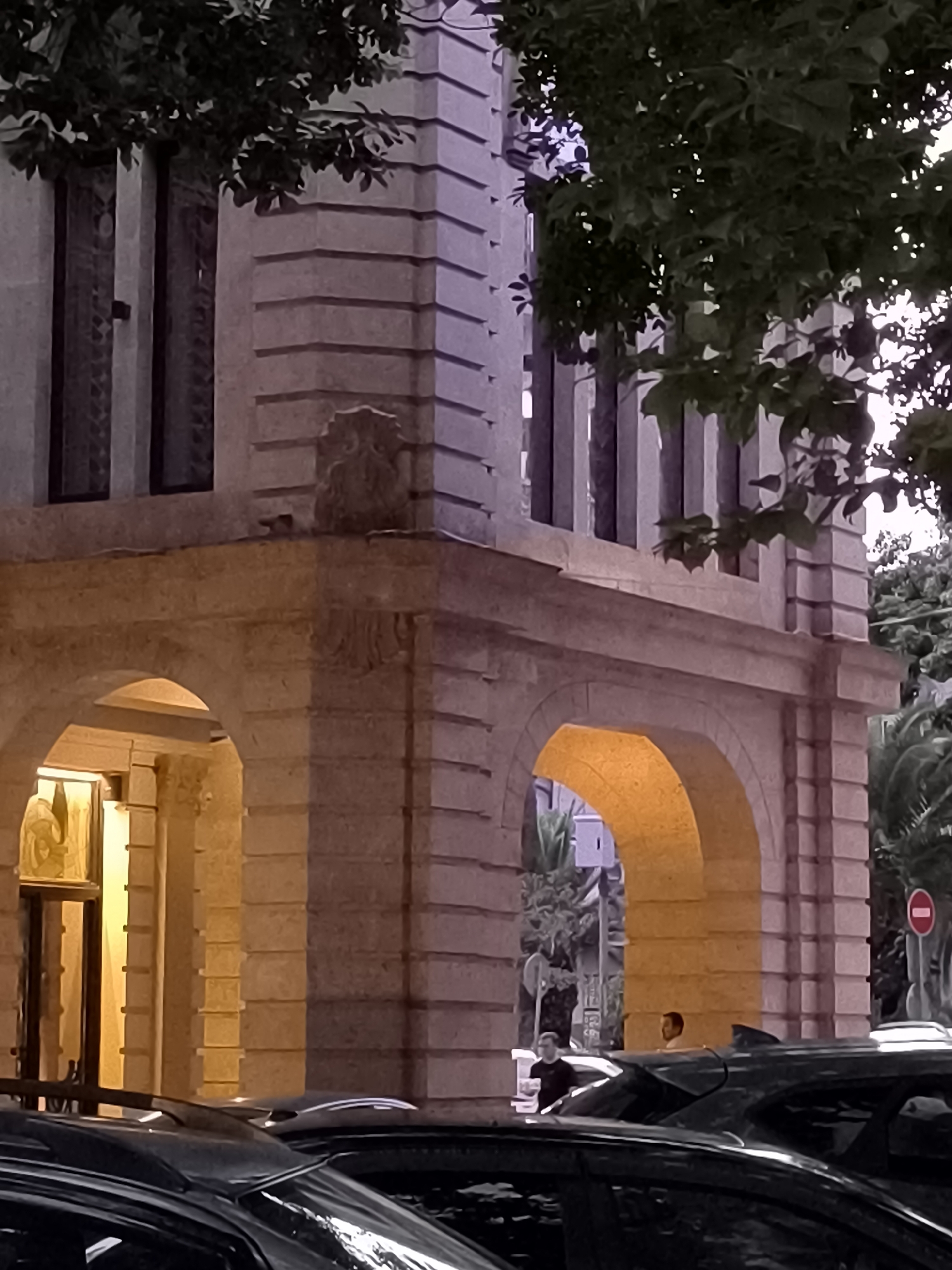 The image shows a street scene in Batumi, Georgia, at dusk. In the foreground, several cars are parked. Behind the cars, a building with arched entrances and stone detailing is visible. A man stands in one of the arches, and another man is seen walking in the background. Trees and foliage are present, with some leaves obscuring the building's upper portion. The lighting suggests artificial illumination from within the building and the street. A red and white circular sign is visible on a pole to the right.