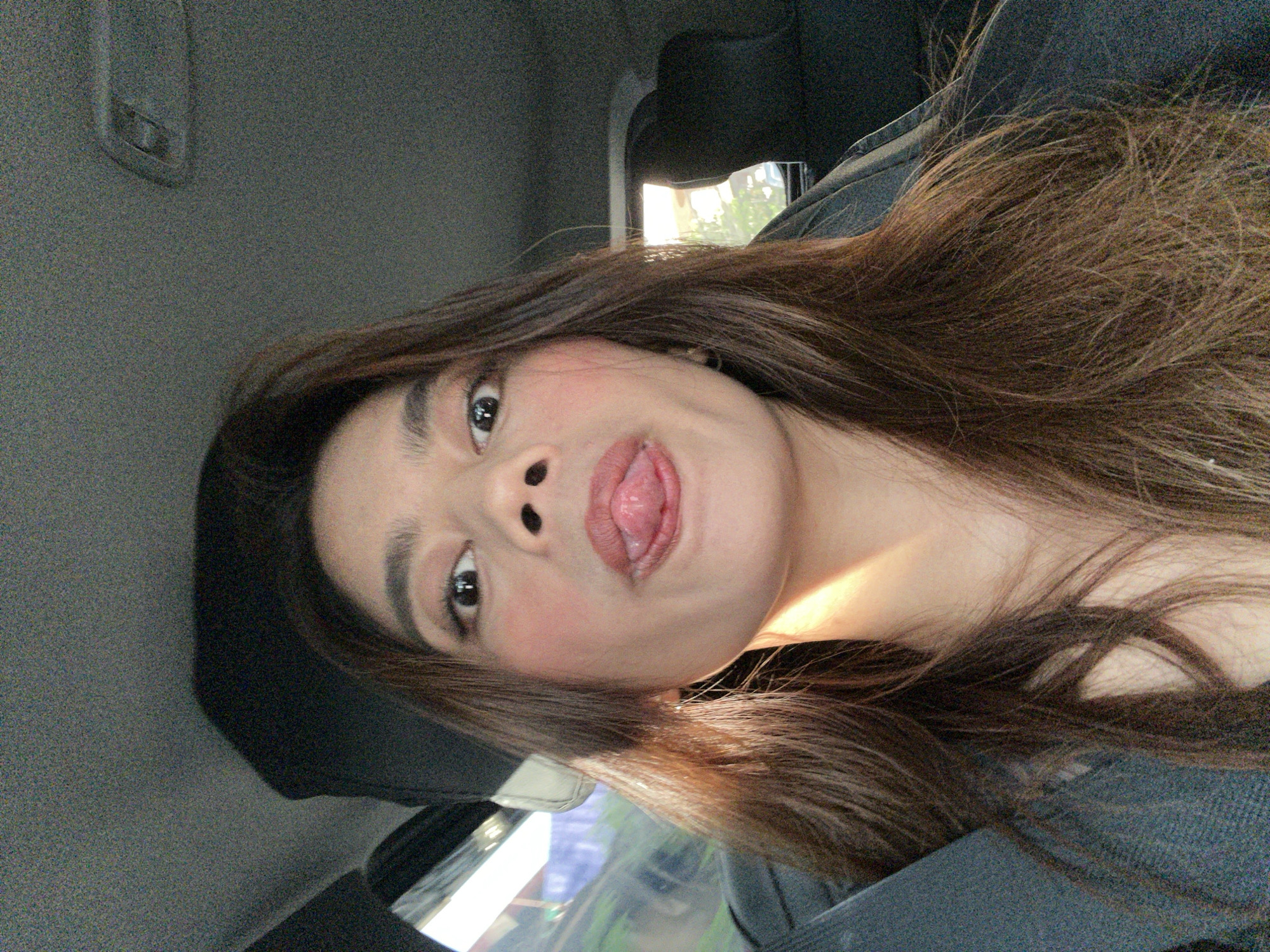 A front-facing portrait captures an adult female occupant inside a vehicle, located in Davao City, Philippines. She is looking directly at the camera with wide, dark eyes and has her tongue playfully extended between her slightly parted lips. Her long, dark brown hair, with lighter streaks, frames her face and falls over her shoulders. She wears a dark-colored garment, and a dark seatbelt strap is visible across her left shoulder. Strong natural light from the left (viewer's right) illuminates her neck and a portion of her hair, creating a bright highlight. The vehicle's interior features a textured gray headliner and dark upholstery on the visible seat backs. Blurred exterior views through the windows reveal green foliage and bright daylight, consistent with a daytime setting.