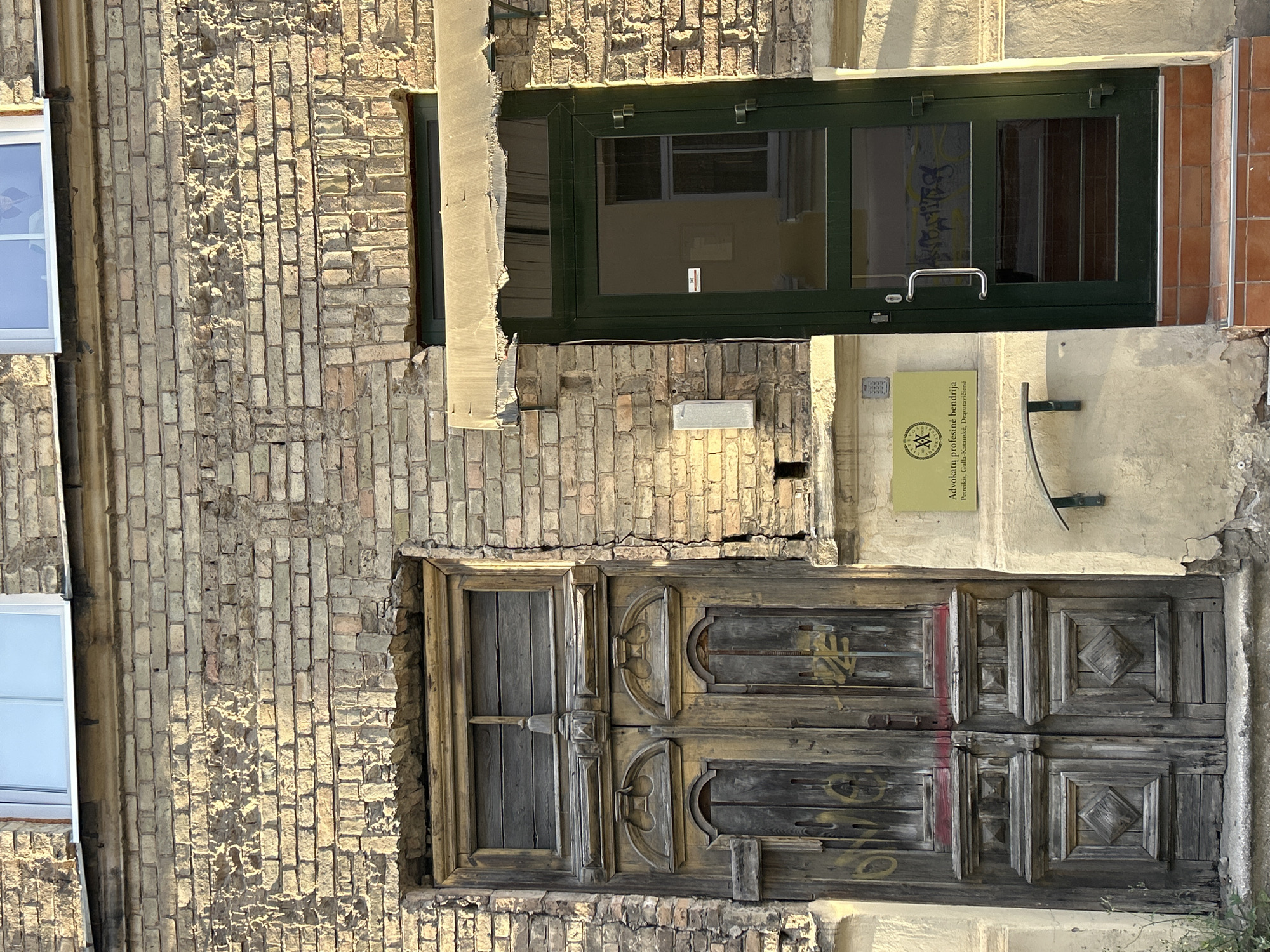 An exterior, eye-level, full shot shows the weathered facade of a building in Vilnius, Lithuania, under natural sunlight. The brickwork is aged and cracked, with a section of the wall appearing to have crumbled near an old, wooden double door on the left. This door, with visible graffiti, exhibits ornate carvings and a single wooden panel window at the top. To its right, a modern, dark green glass door stands open, revealing a dimly lit interior with a staircase visible. Above the doors, two windows with white frames are present. A yellow sign with text and a circular emblem is mounted on the wall between the two doorways. A metal awning, frayed and damaged, is attached above the green door. The ground in front of the entrance is paved with reddish-brown tiles.