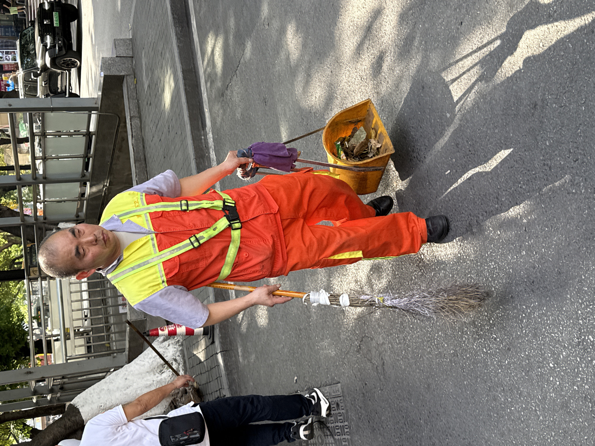 The image, taken in Yuzhong District, China, depicts a street cleaner at work on an urban street or sidewalk during daylight hours.
The primary subject is a man wearing a bright orange jumpsuit with a yellow reflective safety vest over a light-colored short-sleeved shirt, and black shoes. He has a shaved head and a neutral, possibly weary, expression. He is holding a traditional-style broom made of bundled twigs or fibers in one hand, and a long-handled tool with a purple cloth wrapped around it, leading to a yellow, open-top bucket or dustpan filled with litter, in his other hand.
Partially visible in the lower left foreground is another person, seen from the back, wearing a white t-shirt, black pants, and sneakers, carrying a stick.
The setting is an outdoor, paved area, likely a street or busy pedestrian path. The ground is asphalt, and strong sunlight casts distinct shadows, indicating a sunny day. In the background, metal railings suggest a barrier or overpass, and several vehicles, including a black car, are parked or moving. The license plate of the black car is partially visible, showing "渝 A3G9T8," with "渝" being the abbreviation for Chongqing, which includes Yuzhong District. Further back, buildings or structures are visible. A white-painted tree trunk is also present near the bottom left.