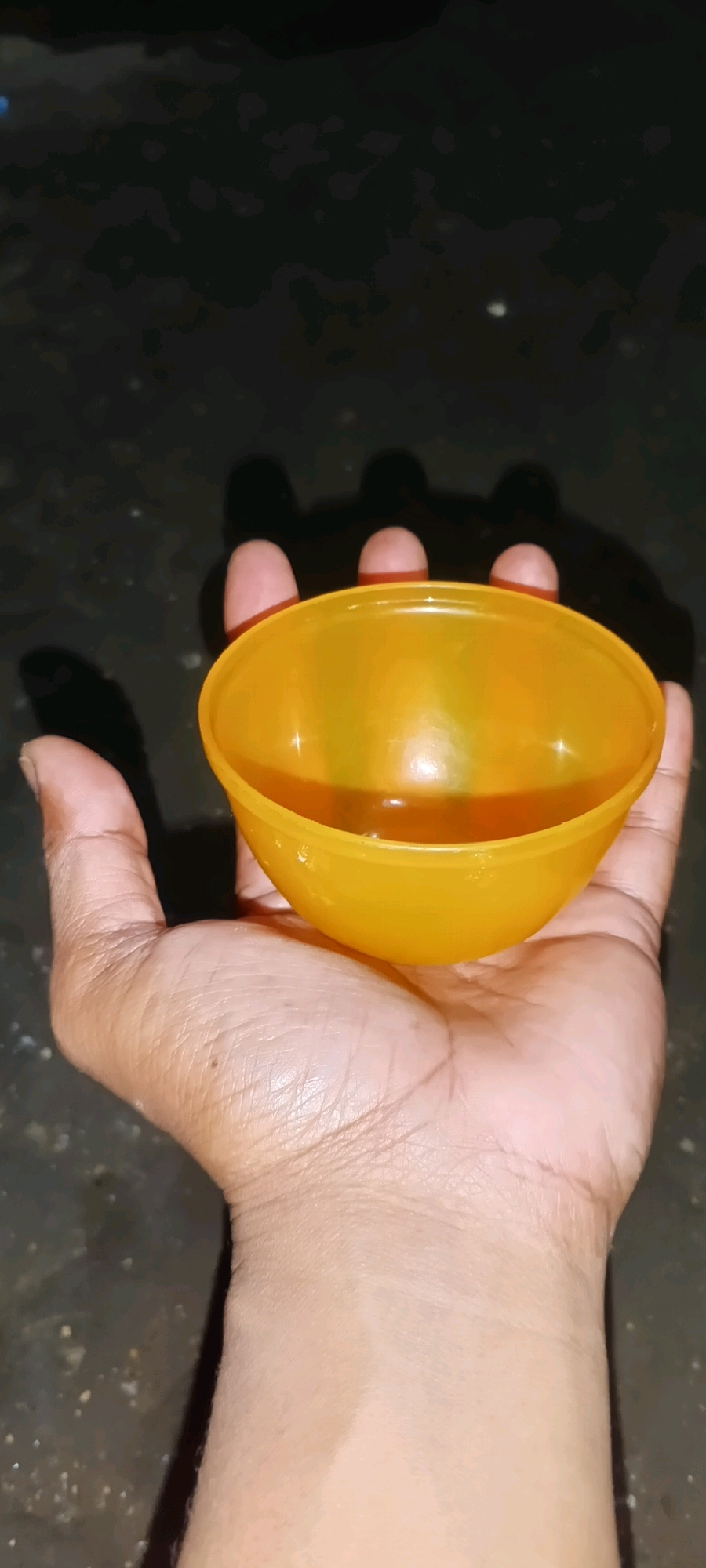 A hand holds an orange bowl containing a small amount of liquid. The background is dark and appears to be a rough, textured surface. The image is shot from a high angle, looking down at the hand and bowl. The lighting is dim, creating shadows on the background and highlighting the glossy surface of the bowl. The subject appears to be indoors or in a shaded outdoor area, possibly in Penanggungan, Indonesia.