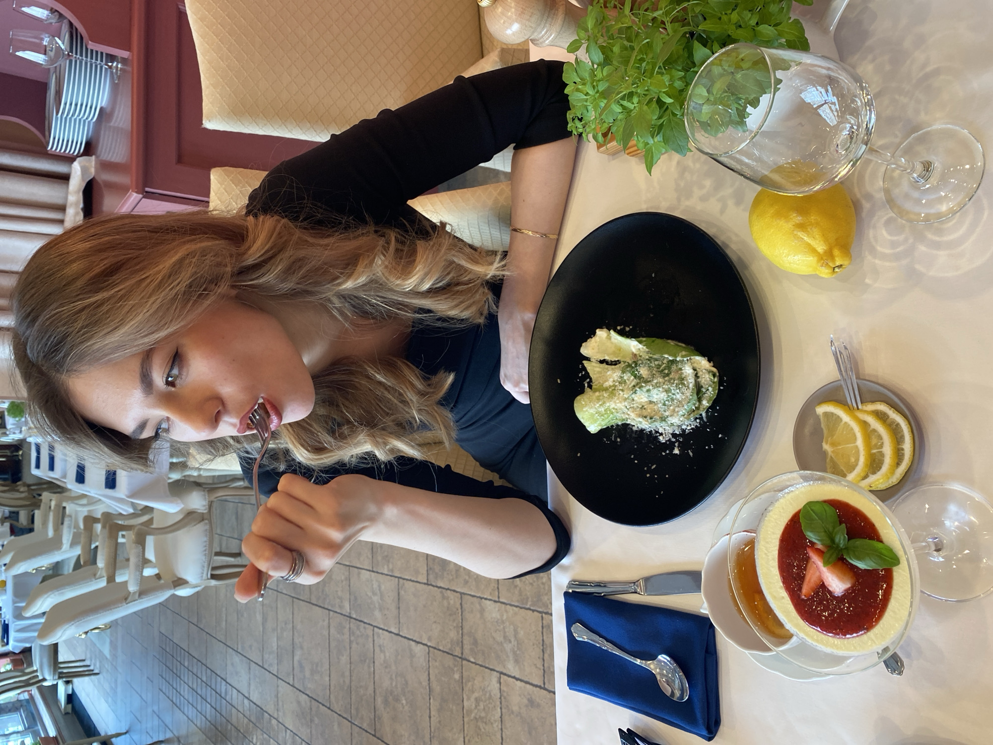The image captures a young woman dining in a restaurant, possibly located in Yekaterinburg, Russia.
**Subjects:** The primary subject is a woman with long, wavy, light brown hair, wearing a black long-sleeved top and a golden bracelet on her right wrist. Other subjects include various food items, an empty wine glass, cutlery, a blue napkin, a whole lemon, lemon slices, and a potted green plant. The background features restaurant furniture like other tables, chairs, and wall paneling.
**Activity or event taking place:** The woman is actively eating a meal. She is shown in the act of bringing a forkful of food towards her mouth, indicating she is mid-meal or enjoying her dish. It appears to be a casual dining experience in an indoor setting.
**Notable details:**
* **Food:** On a black plate, there is a green salad, possibly a Caesar salad, topped generously with grated white cheese. Adjacent to it, a dessert in a glass cup features a light-colored creamy base topped with a vibrant red strawberry sauce, garnished with a fresh strawberry slice and a few basil leaves. A small dish containing a golden liquid, likely honey or syrup, accompanies the dessert, along with a separate small plate holding three slices of lemon.
* **Table setting:** The table is covered with a white tablecloth and features standard dining cutlery including a knife, a spoon resting on a folded blue napkin, and the fork the woman is using. An empty stemmed wine glass and a whole fresh lemon are also present. A potted plant with green leaves, possibly an herb like basil, serves as a decorative element.
* **Setting:** The restaurant interior is well-lit, suggesting daytime. The background shows light-colored cushioned chairs and tables with white tablecloths, along with warm-toned wooden wall paneling and a tiled floor.
* **Emotion:** The woman appears focused on her meal.
* **Visible text:** There is no visible text in the image.