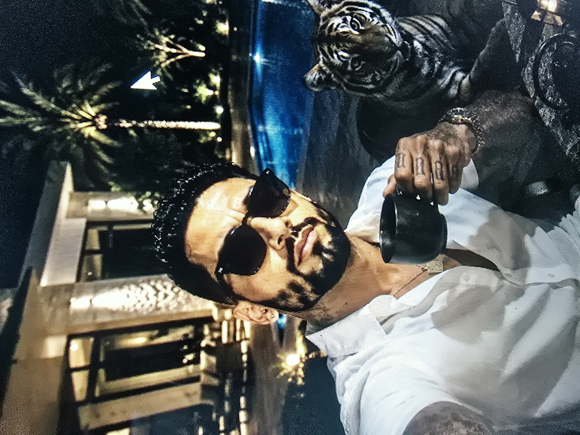 A man wearing a white collared shirt and sunglasses is taking a selfie with a tiger in an outdoor setting at night. The man is holding a black mug in his left hand. He has tattoos on his fingers and a bracelet on his wrist. The tiger is lying down next to him. In the background, there is a swimming pool and palm trees. There is also a building visible in the distance. The scene appears to be a residential area.