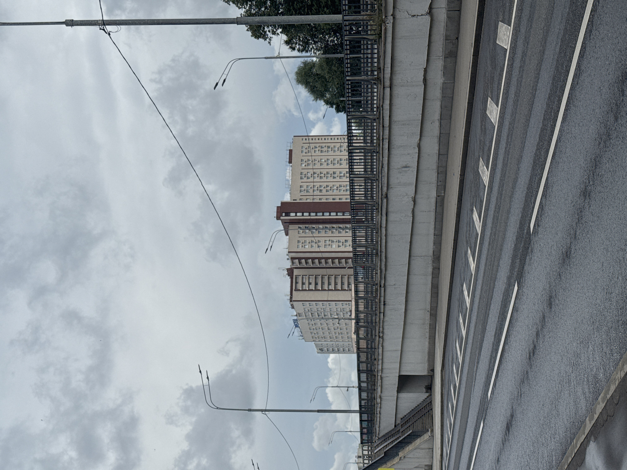 The image depicts an overcast day in Moscow, Russia, featuring a multistory apartment building in the background. The building is light-colored with darker reddish-brown accents on its facade. In the foreground, there is a multi-lane road with white lane markings and some wet patches, suggesting recent rain. A concrete overpass with a metal railing runs horizontally across the mid-ground, separating the road from the building. Stairs leading up to the overpass are visible on the left side. Several streetlights and power lines are present throughout the scene. Trees are also visible behind the overpass on the right side of the image.