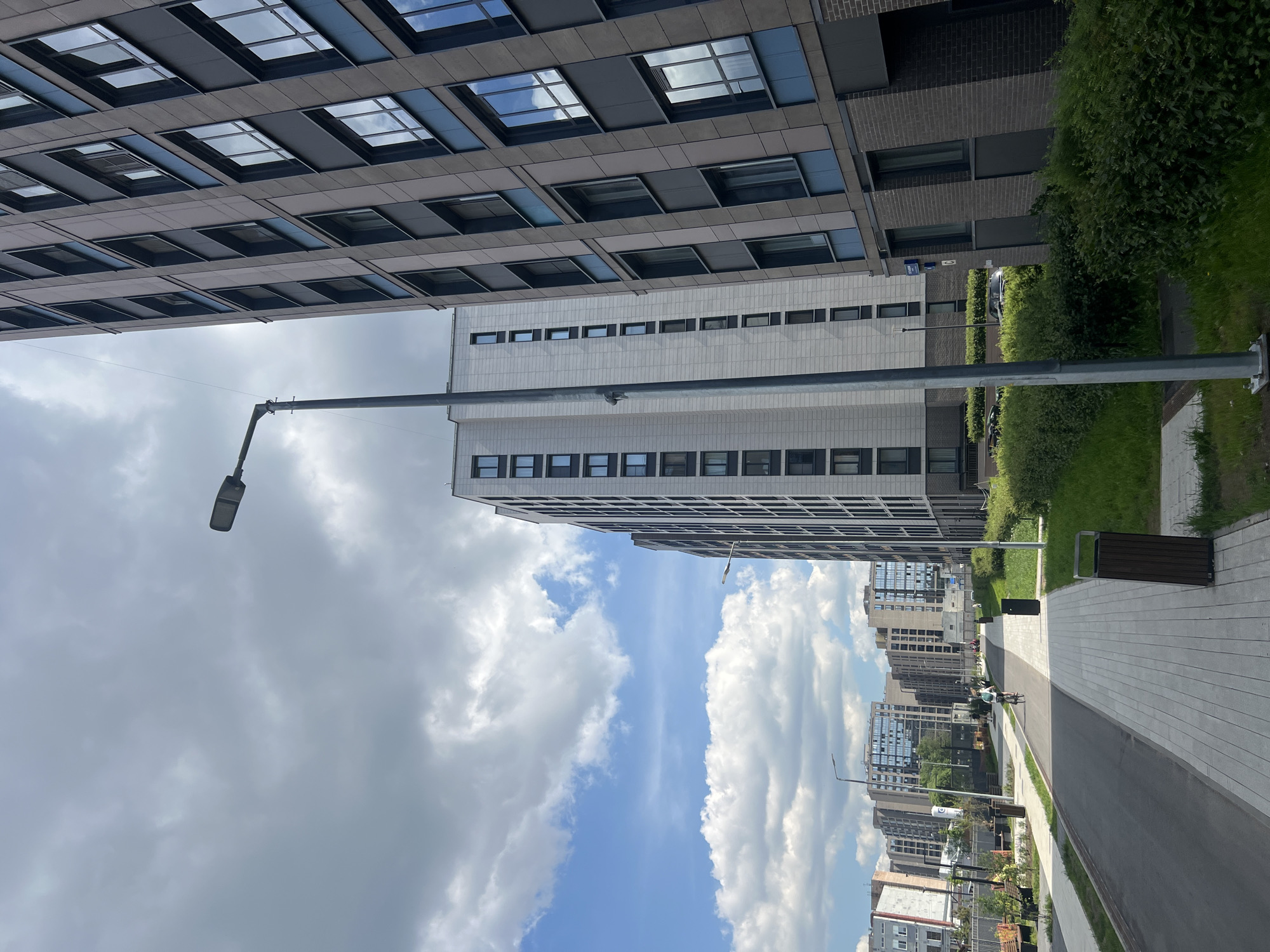 The image shows a street scene in a modern residential area in Saint Petersburg, Russia. In the foreground, a paved walkway runs alongside a road. A person is cycling on the road. Several tall, contemporary apartment buildings with numerous windows line the street. Greenery, including grass and bushes, is visible between the buildings and along the walkway. A street lamp stands on the right side of the walkway. The sky is partly cloudy with some blue sky visible.