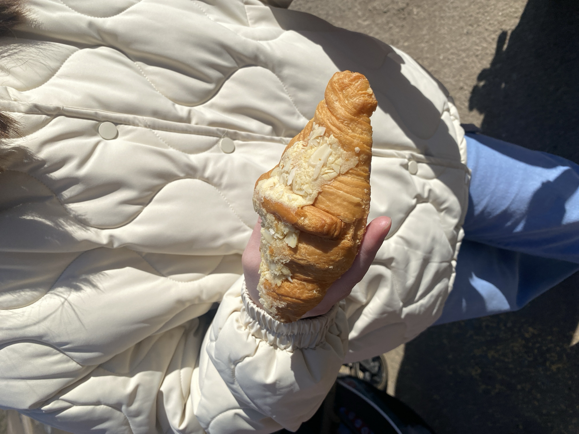A person is holding a croissant covered with sliced almonds in their right hand. They are wearing a white puffer jacket and a light blue shirt. The background is blurred and shows a concrete surface. The location is uncertain, and there is no verifiable connection to Chaikovsky, Russia.