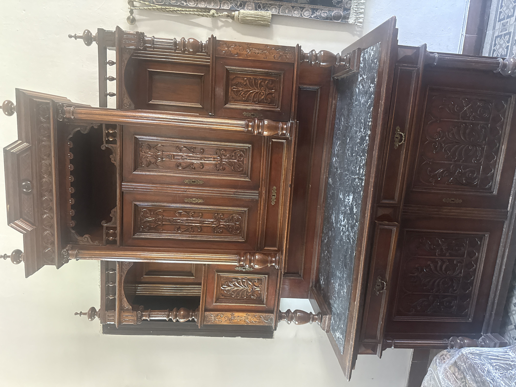 A close-up shot features an antique wooden sideboard with intricate carvings and a marble top. The sideboard has multiple compartments, including drawers and cabinet doors, some of which are open. The photograph is taken indoors, and the background shows a plain wall. The top of a rug or tapestry with tassels is visible in the background. The location is not discernible from the image. No people are present in the image. The city of Karpaty, Ukraine, is not identifiable from the image.