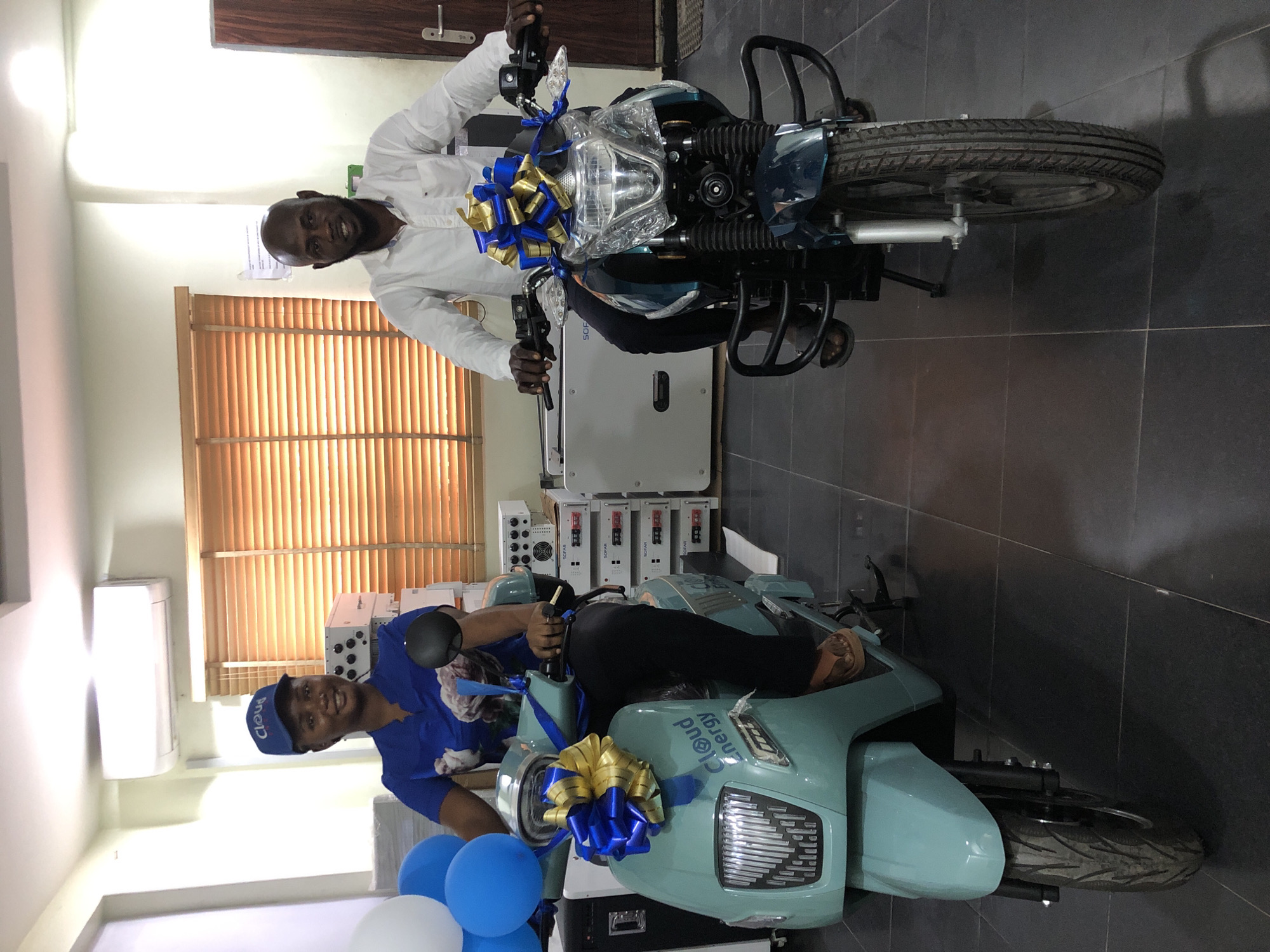 In Ikeja, Nigeria, a woman wearing a blue cap and a blue top with a floral pattern sits on a light blue electric scooter. She is smiling and holding a black circular object in her right hand. To her right, a man in a white shirt is seated on a dark teal motorcycle. Both vehicles are adorned with large blue and gold bows. In the background, stacks of white electronic equipment, possibly power inverters, are visible, along with blinds on a window and a door. The floor is tiled with dark gray squares.