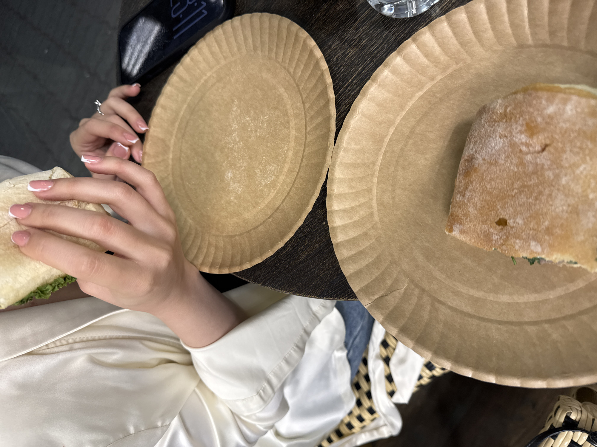 From an overhead perspective, the image captures a person holding a sandwich with lettuce inside. The person is wearing a white silk shirt and blue jeans with a straw bag lying beside them. The nails are well-manicured with white tips and a ring is visible on one of the fingers. On the wooden table are two paper plates, one empty and the other holding a half-eaten sandwich. A black mobile phone is located on the table. The location appears to be Yerevan, Armenia.
