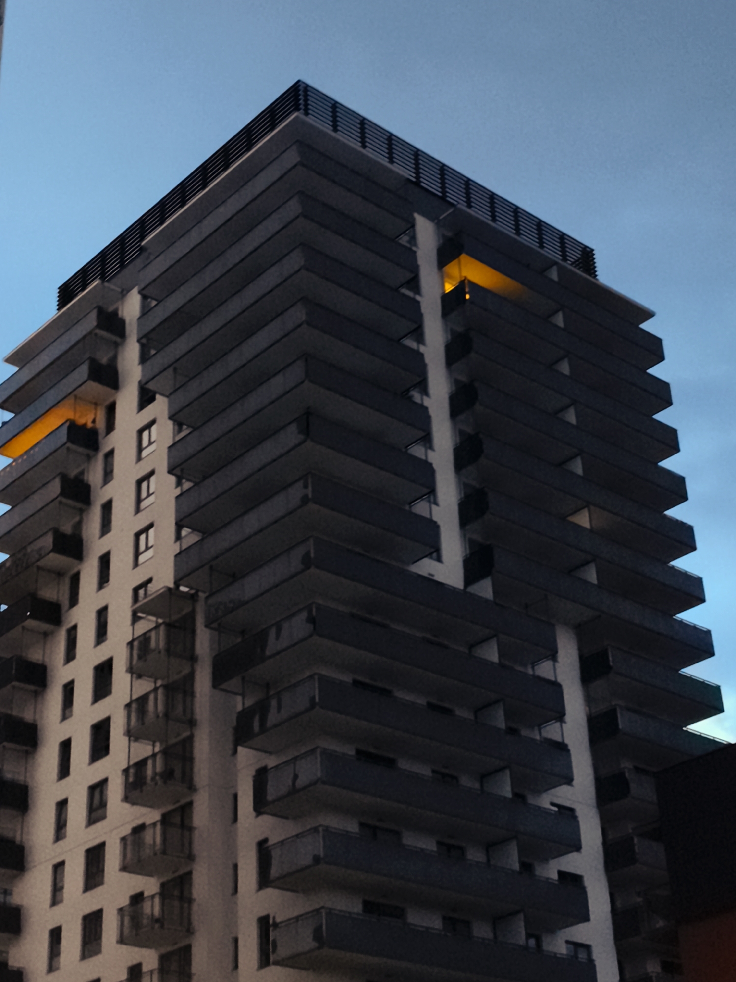 A low-angle, vertical shot captures a modern apartment building against a twilight sky. The building features a repeating pattern of dark grey balconies jutting out from a lighter facade. Several windows are visible on the lighter sections of the building. One balcony on the upper floors glows with a warm, orange light, suggesting interior illumination. The building is topped with a dark railing. This building appears to be located in Gdańsk, Poland, specifically in the vicinity of the former shipyard area, characterized by its contemporary architectural style. The overall atmosphere is calm, with the city lights beginning to emerge in the fading daylight.