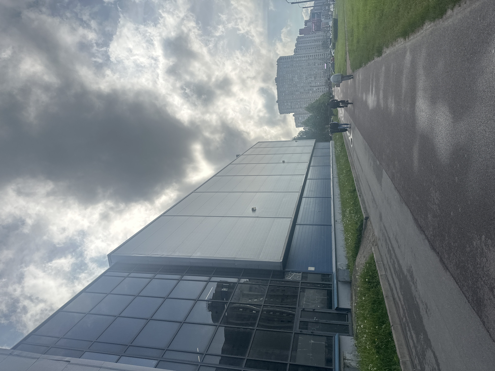 An overcast day in an urban setting. A modern building with large glass windows and a grey metal facade dominates the left side of the frame. A blue lower section runs along the base of the building. In front of the building is a paved walkway with a green lawn to the right. Several people are visible on the walkway: one person on a bicycle, and three people walking. In the background, large apartment buildings stretch across the horizon under a dramatic, cloudy sky. Cars are parked on the right side of the image. The overall scene depicts a utilitarian, modern urban environment on a cloudy day.