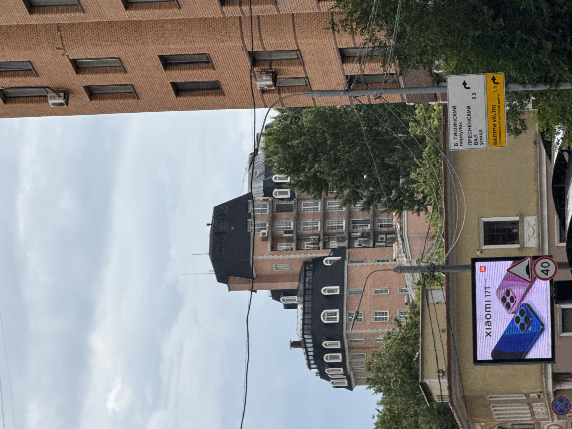 The image shows a street scene in Moscow, Russia, on a cloudy day. In the foreground, a large digital billboard displays an advertisement for "Xiaomi 17T," featuring two smartphones. To the right of the billboard, a street sign points towards "Б. ТИШИНСКИЙ переулок," "ПРЕСНЕНСКИЙ ВАЛ улица," and "ВАЛТРИ VÄLTRI премиальный деловой центр." A speed limit sign indicating "40" is visible below the street sign.
Dominating the background are several multi-story buildings. The building on the far right is a modern, brick-clad structure with many windows and air conditioning units. To its left are two older buildings with distinctive mansard roofs, characteristic of St. Petersburg architecture, but also found in Moscow. One of these buildings has a rounded facade with numerous arched windows. Green trees are interspersed throughout the scene, suggesting a relatively urban but leafy environment. Overhead, power lines crisscross the sky. The overall impression is of a bustling city street with a mix of modern and older architecture.