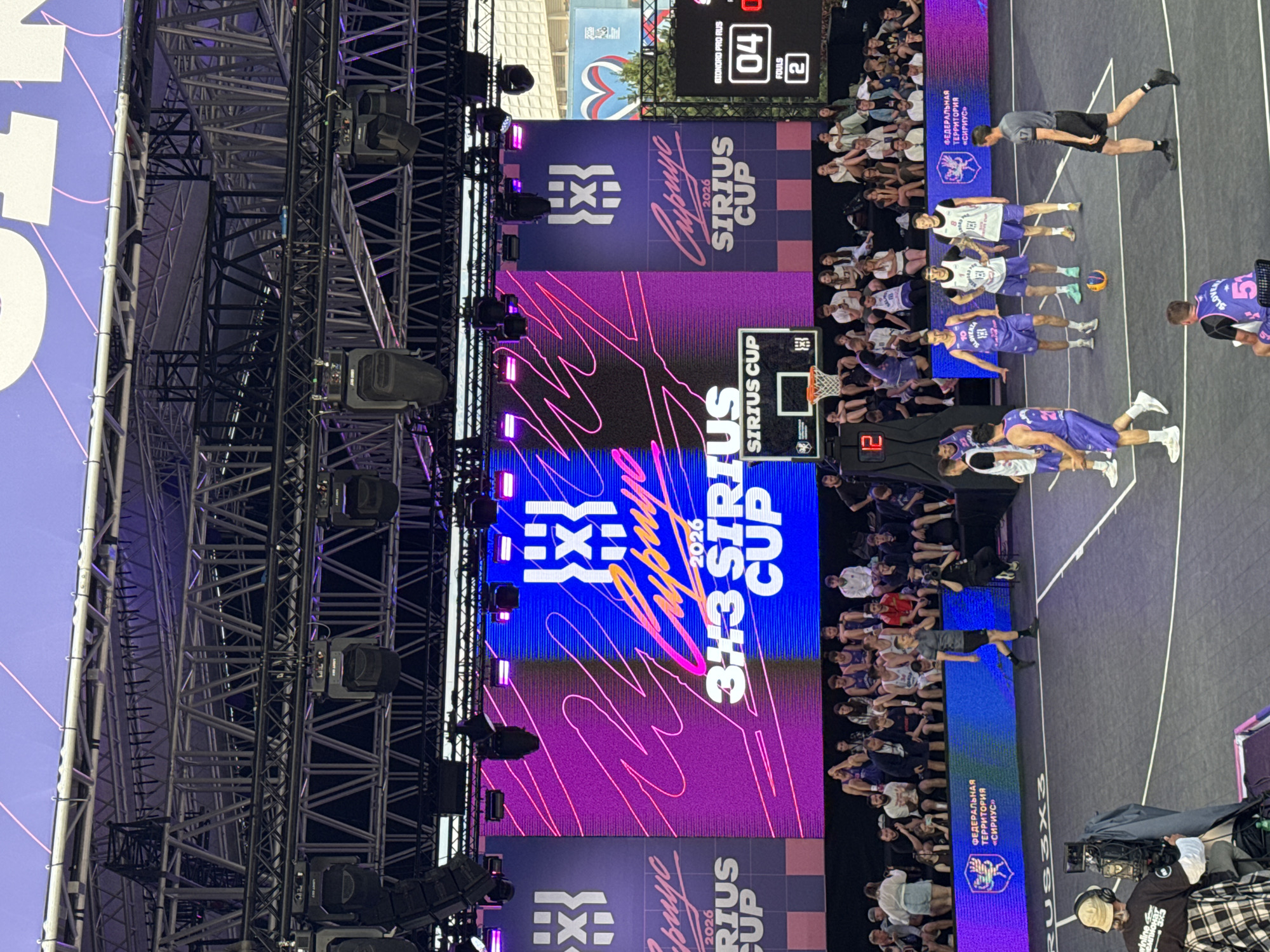 This image captures an outdoor 3x3 basketball tournament taking place in Sirius, Russia. The scene is set at a court with a large digital screen displaying event graphics and a score. Spectators are seated in bleachers behind the court, watching the game. Several players, dressed in team uniforms, are on the court, some in active play and others observing. A referee in uniform is also present on the court. The background shows a large stage structure with lighting equipment, suggesting a professional event setup. The overall atmosphere appears to be one of a sporting competition with an engaged audience.