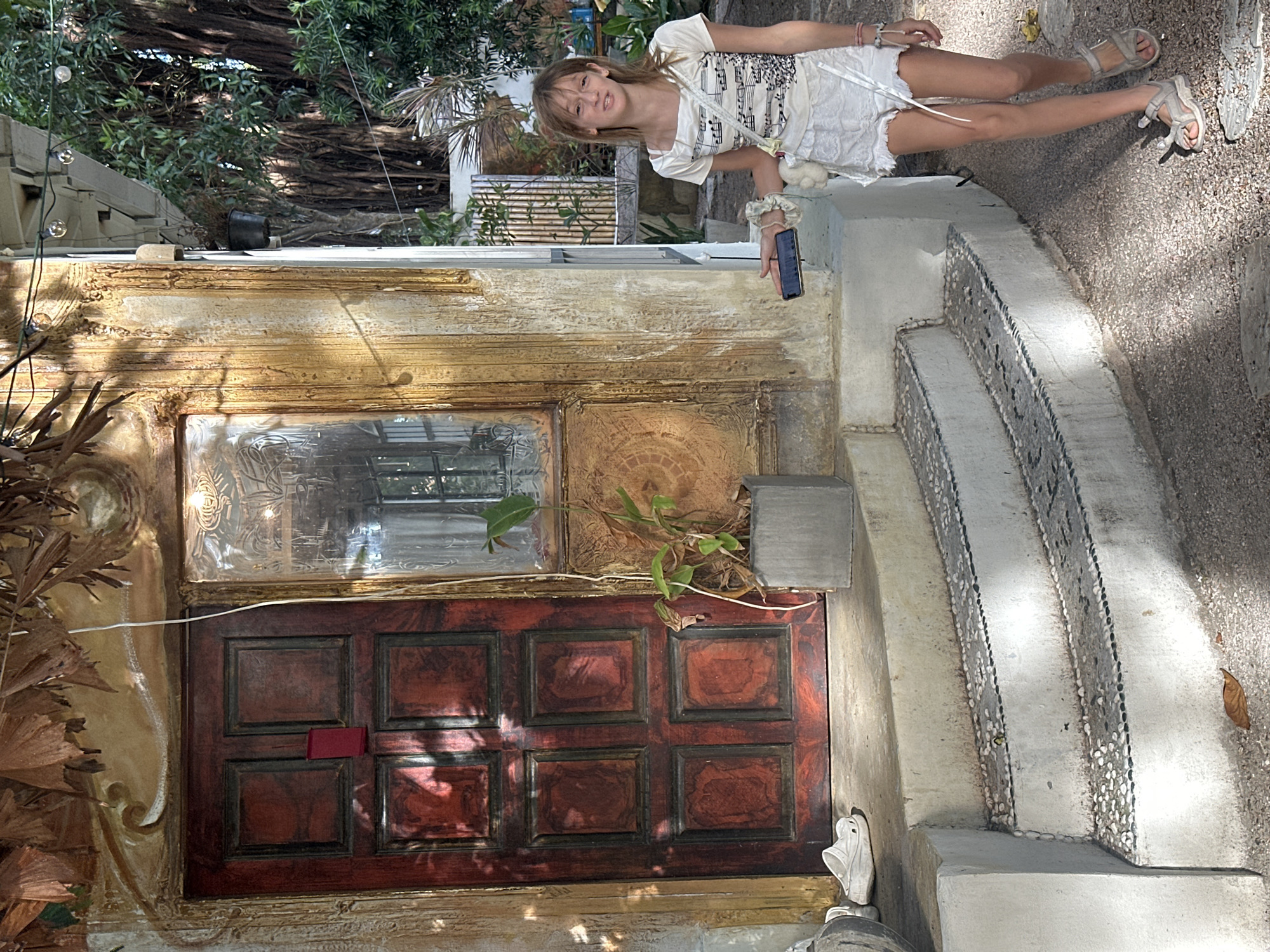 A young woman with blonde hair is posing for a photo in an outdoor setting. She is wearing a white t-shirt with a musical score print and a white skirt. She has a small white fluffy bag hanging from her shoulder and is holding a smartphone in her right hand. The woman is standing on a textured stone pathway next to a building with an ornate, aged wooden door and a decorative window. The door is a rich, dark red with a grid of six panels. Above the door is a rectangular window with a textured glass surface. The building's facade is a creamy yellow with distressed paint and intricate carvings. Green foliage, including palm fronds, is visible around the building. The pathway leads up a set of curved stairs decorated with small pebbles. The time of day appears to be daytime, with sunlight casting shadows. The location is likely a tropical environment, suggested by the lush greenery and warm atmosphere. The woman's expression is relaxed and friendly. There is no visible text in the image.