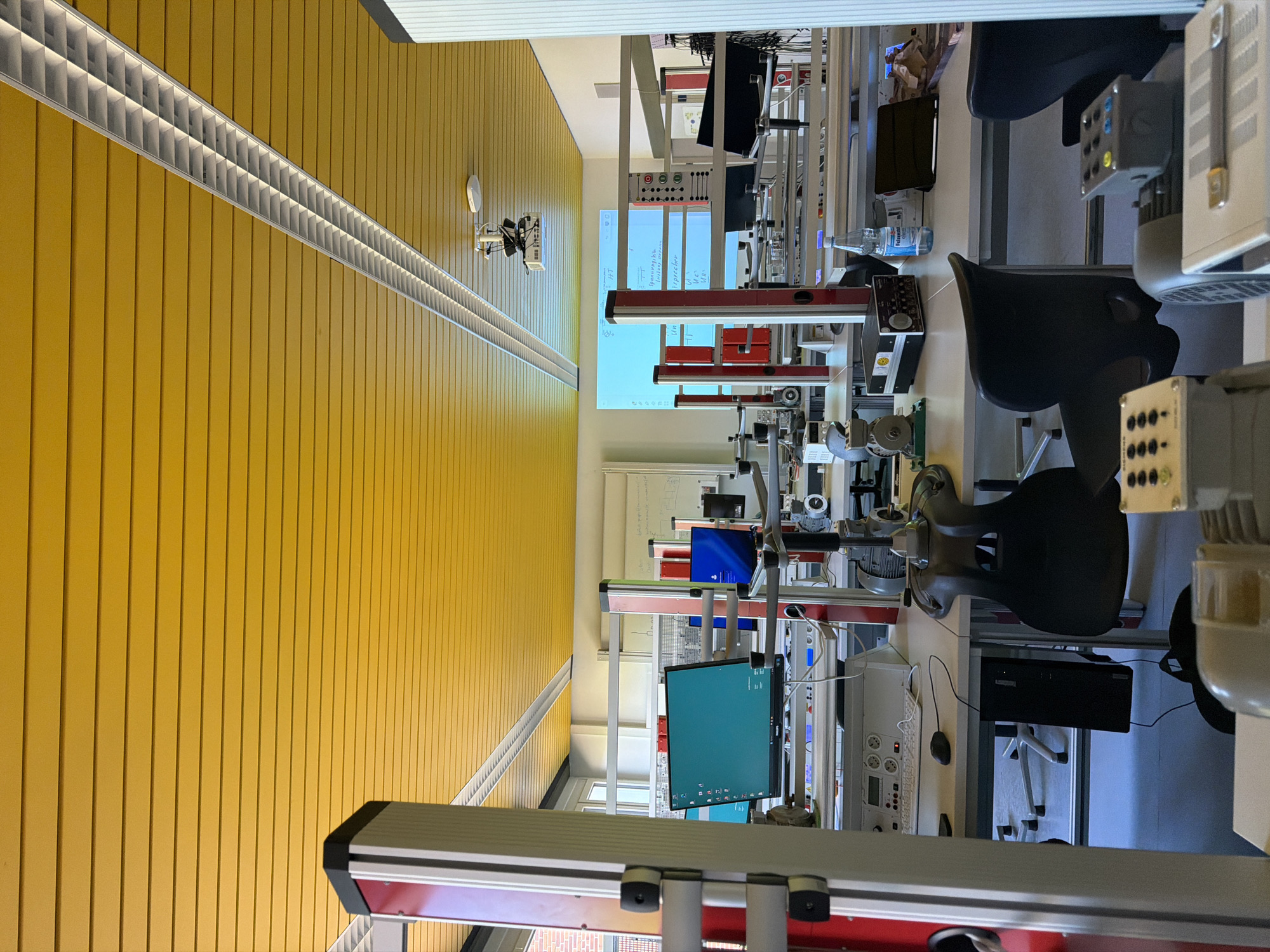 This image depicts a classroom or laboratory setting, likely within a vocational school or university in Warendorf, Germany, given the presence of technical equipment and educational materials. The room features a bright yellow, slatted ceiling with fluorescent lighting fixtures. Several workstations are visible, each equipped with computers, monitors displaying various interfaces, and scientific or technical apparatus, including motors and control panels. Desks are organized in rows, with some featuring chairs. A projector is mounted on the ceiling, pointing towards a projection screen displaying text and diagrams. The overall environment appears organized and functional, geared towards practical learning and experimentation. There are no individuals visible in the image.