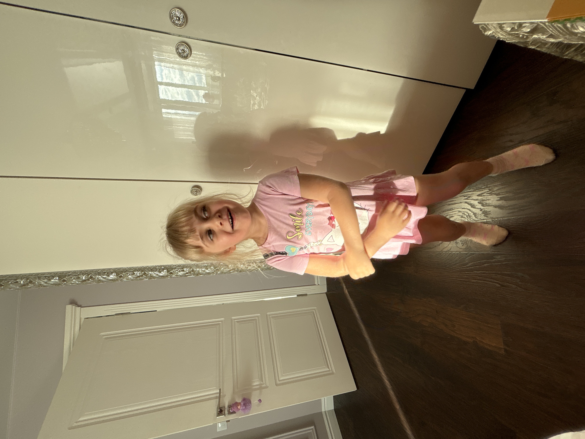 In an indoor scene, a young Caucasian girl with blonde hair stands facing slightly left, with her head turned to look to her right. She is wearing a pink t-shirt with a graphic design that includes the words "Smile Everyday" and an illustration of a cat, and a pink skirt. Her arms are crossed in front of her chest, and she is wearing light pink socks. The background includes a white door on the left, a tall white wardrobe to the right, and a dark wooden floor. Sunlight streams in from the right, casting a shadow of the girl on the wardrobe. The overall lighting is warm and natural.